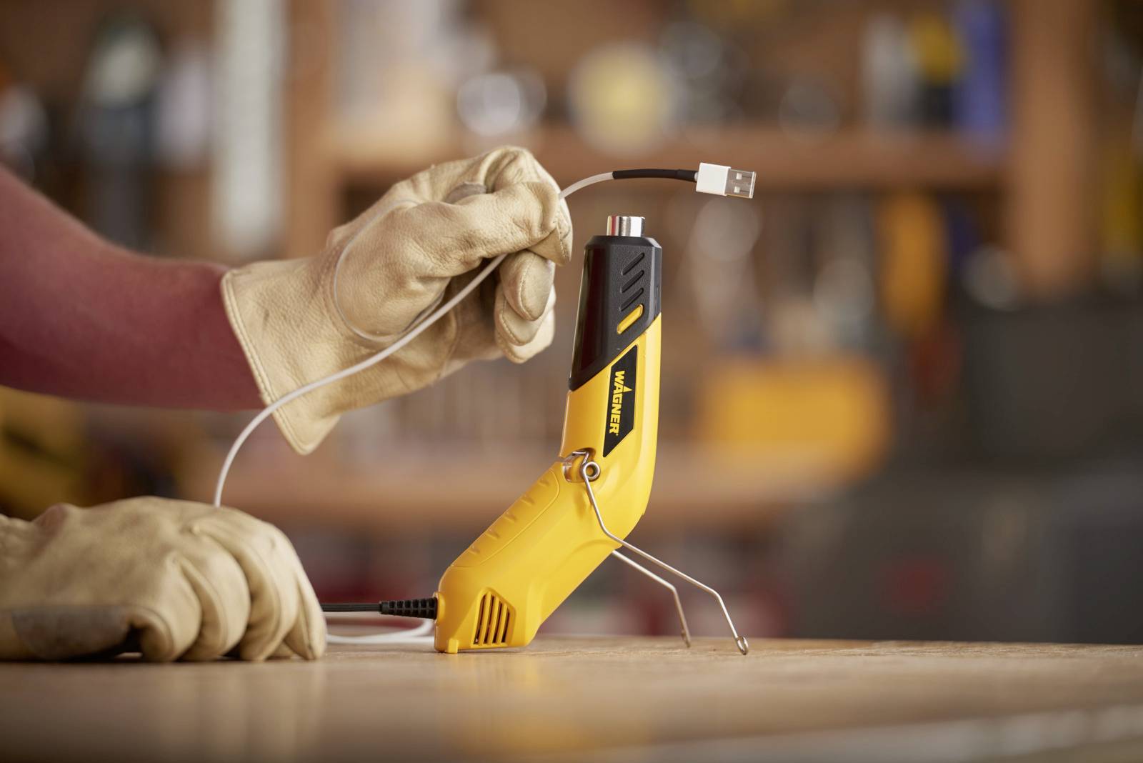 Eine Person mit Arbeitshandschuhen hält ein USB-Kabel über einem gelben Multifunktionswerkzeug in einer Werkstatt.