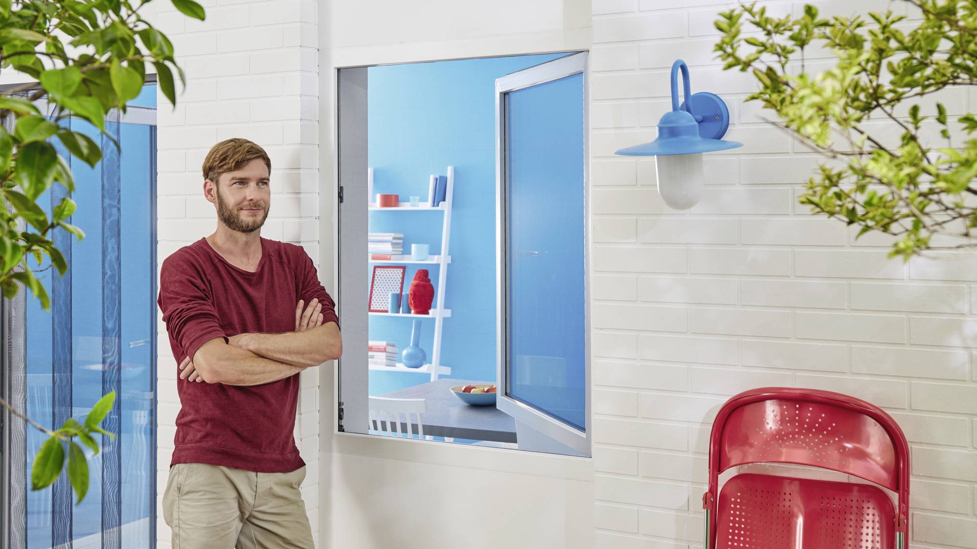 Ein Mann mit rotem Pullover steht vor einem weißen Gebäude mit blauen Details, ein roter Stuhl im Vordergrund, Bäume rahmen die Szene.