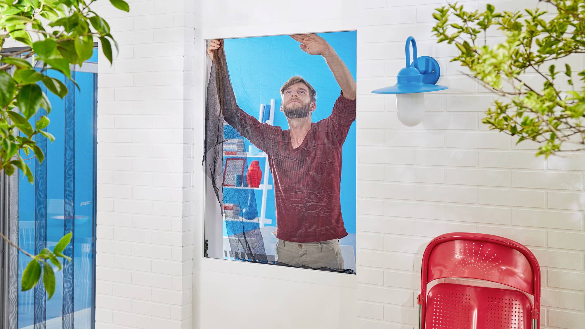 Mann zieht ein Moskitonetz an einem Fenster auf; im Hintergrund blaue Wand und Regal mit roten Gegenständen; sonniger Tag.