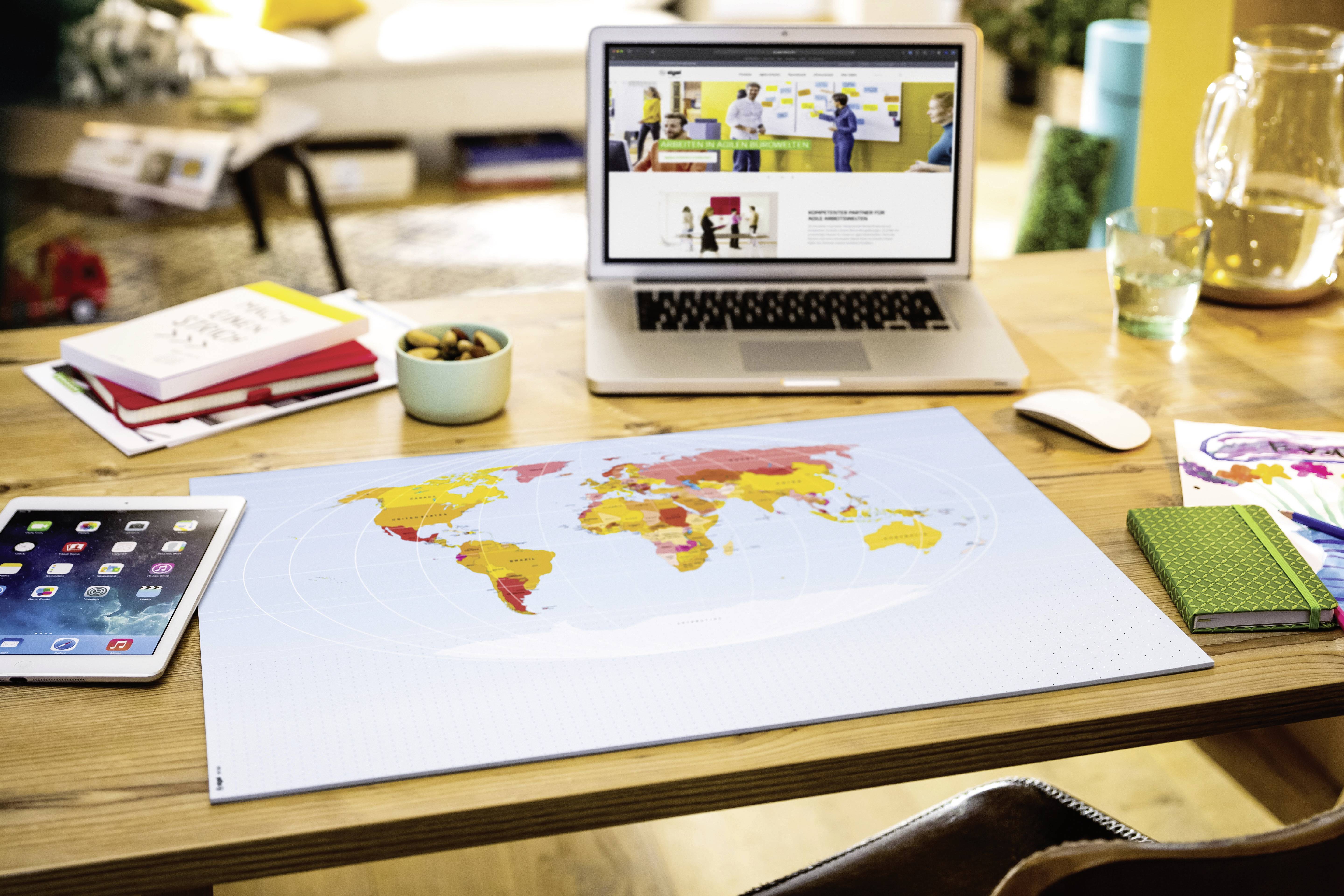 Auf einem Schreibtisch liegen ein Laptop mit einer Website auf dem Bildschirm, ein Tablet, ein Weltkartenposter und Notizbücher.