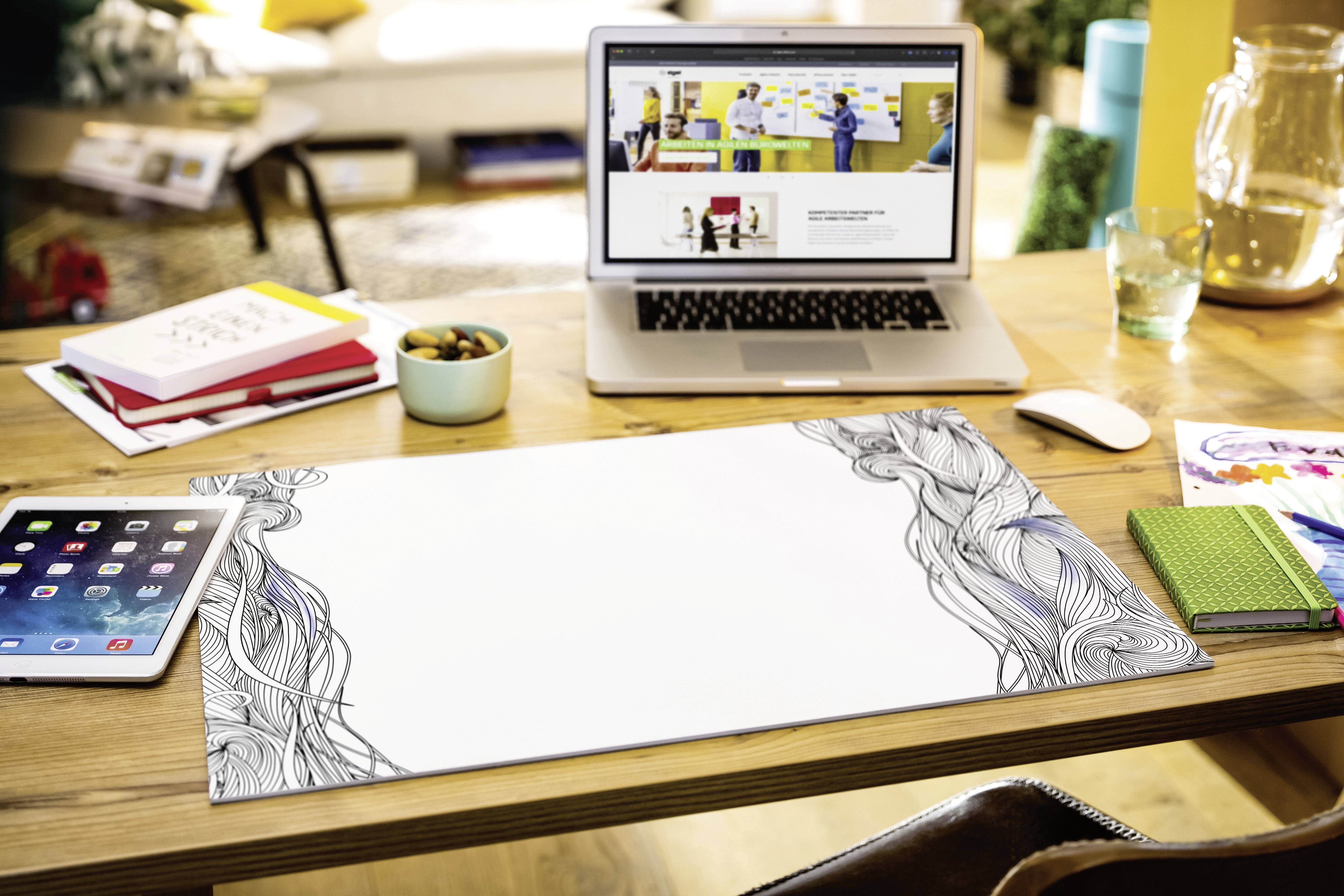 Ein Schreibtisch mit einem Laptop, der eine Website zeigt, einem Tablet, Notizbüchern und einem weißen Papier mit schwarzem Randmuster.