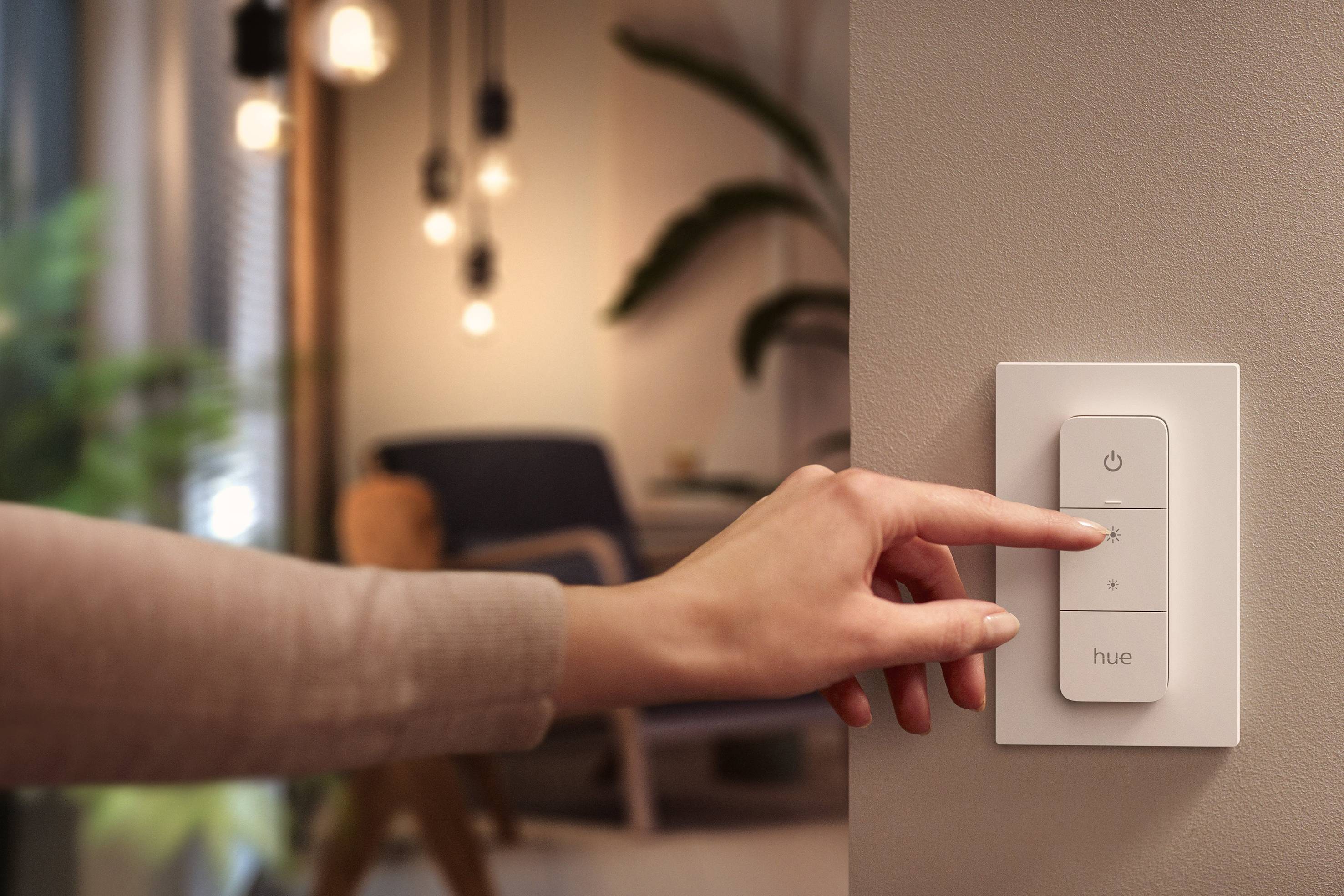 Eine Hand drückt auf einen smarten Lichtschalter an der Wand, im Hintergrund ein gemütlich beleuchteter Raum mit Pflanzen und Stuhl.