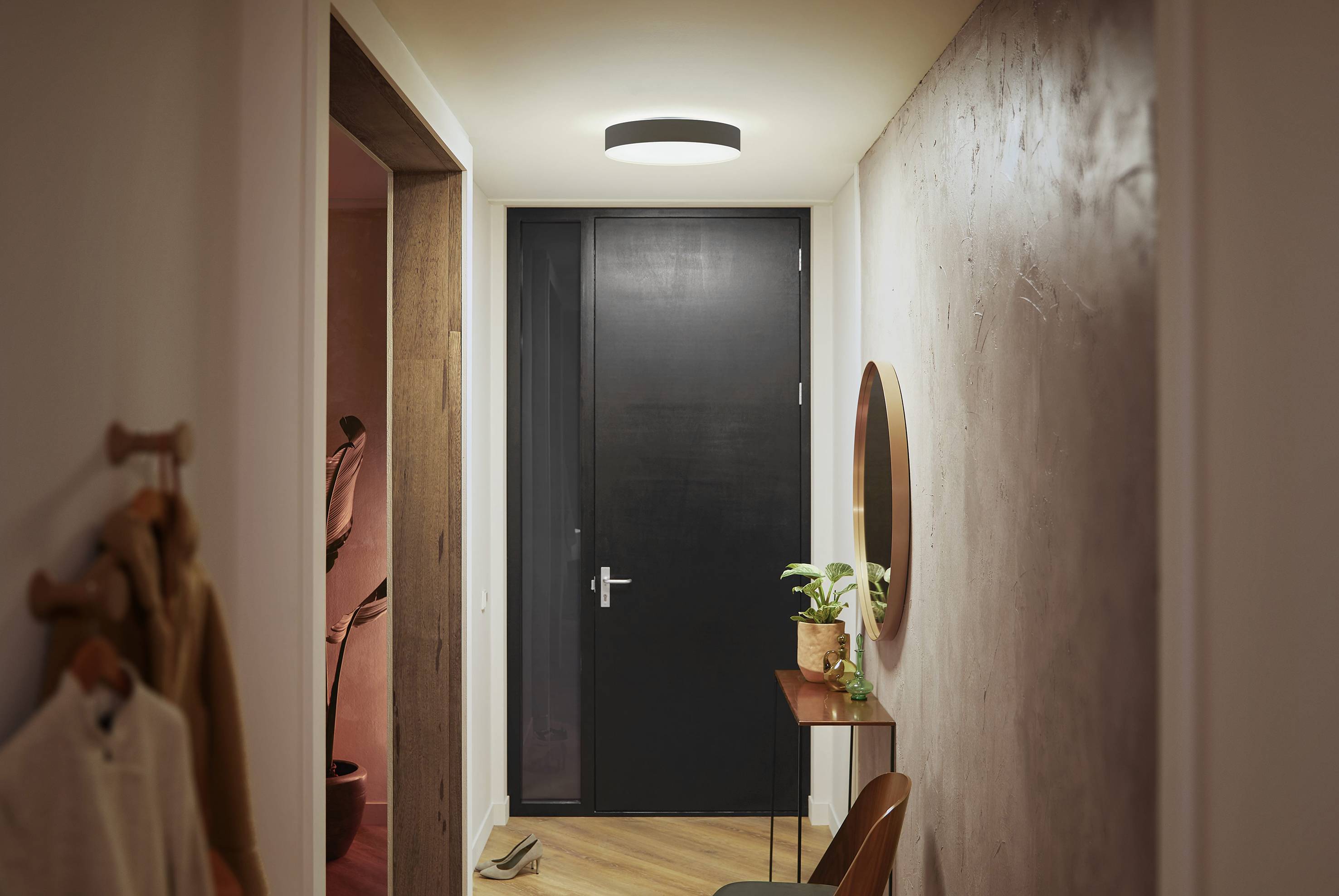 Flur mit heller Deckenleuchte, schwarzer Tür am Ende, kleinem Tisch mit Pflanze und rundem Spiegel an der Wand.