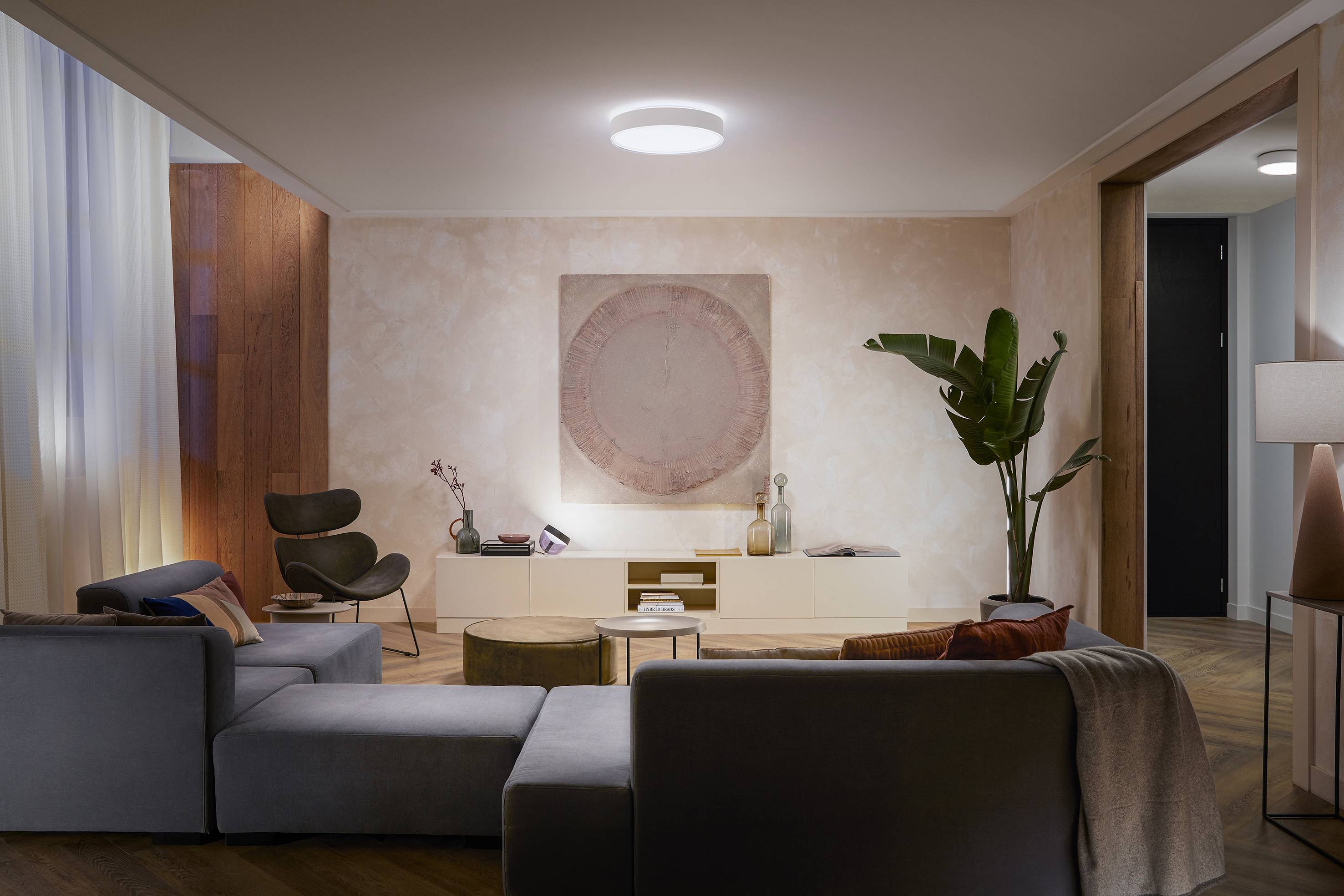 Ein gemütliches Wohnzimmer mit grauem Sofa, Pflanzen und moderner Beleuchtung. Ein Gemälde hängt über einem weißen Sideboard.