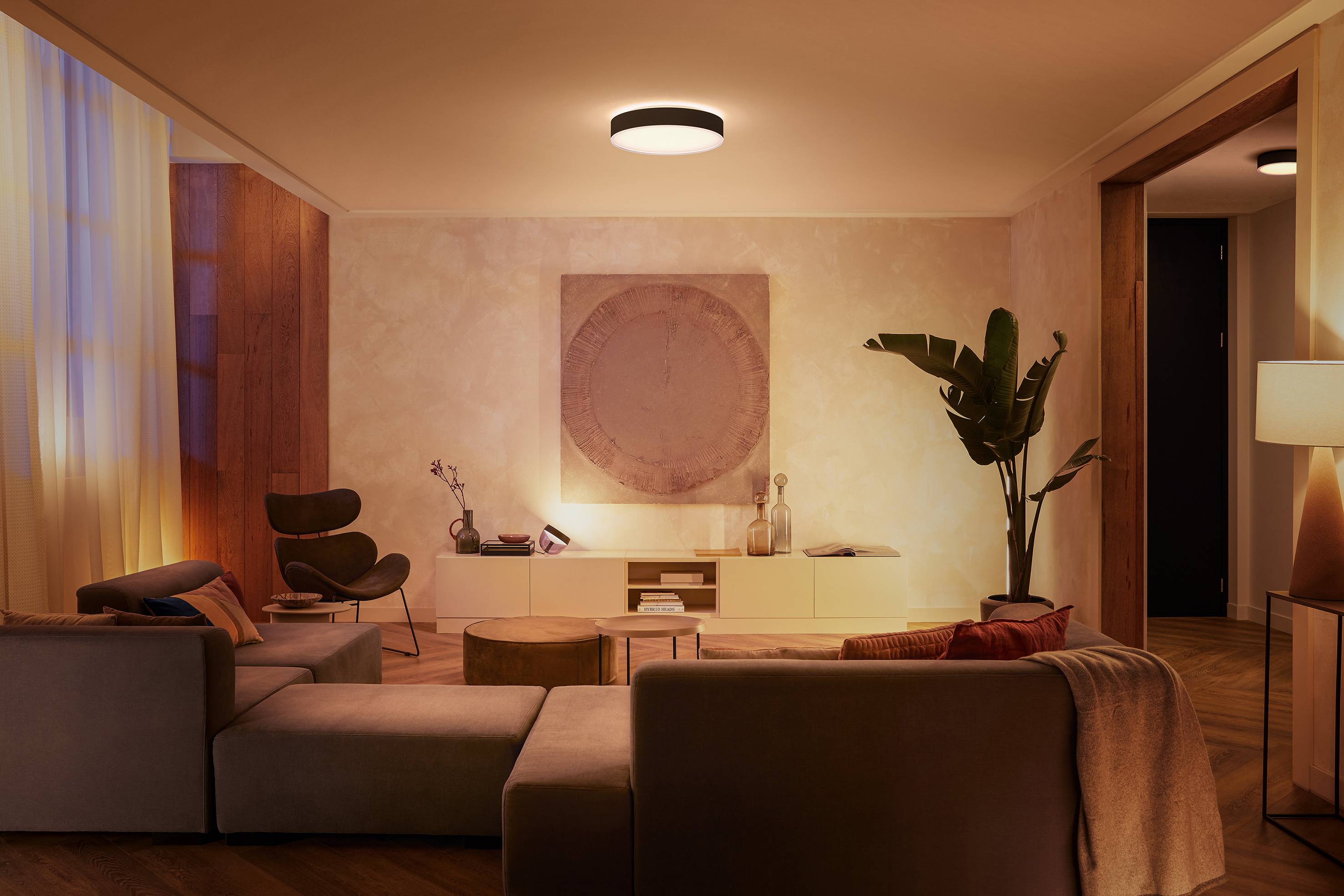 Ein gemütliches Wohnzimmer mit modernen Möbeln, warmem Licht, einem großen Sofa, einem Sessel, einem runden Tisch und Pflanzen.