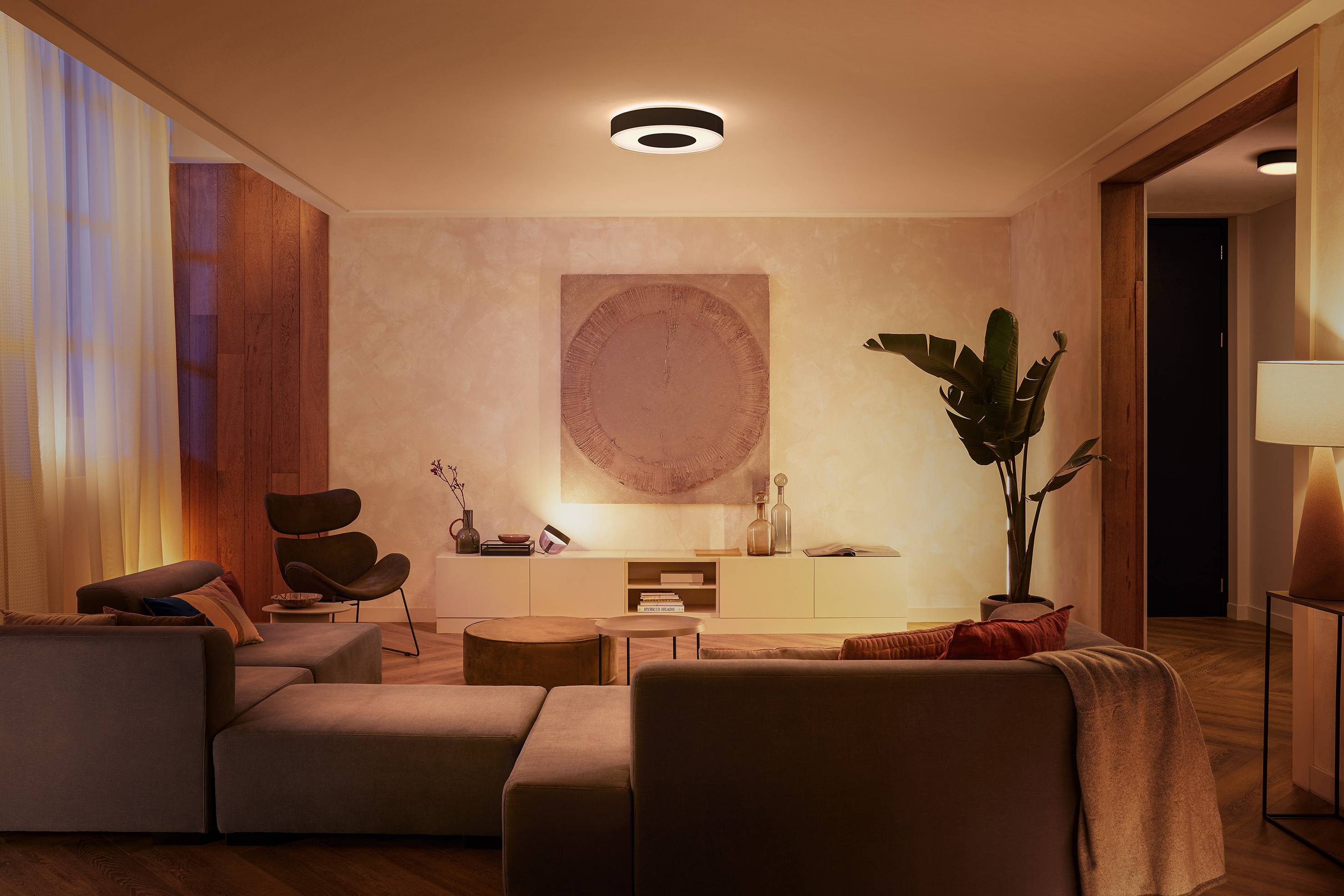 Ein modernes Wohnzimmer mit gedämpfter Beleuchtung, einem Sofa, einem Sessel und einem großen Pflanzenarrangement. Eine Lampe beleuchtet den Raum von der Decke.
