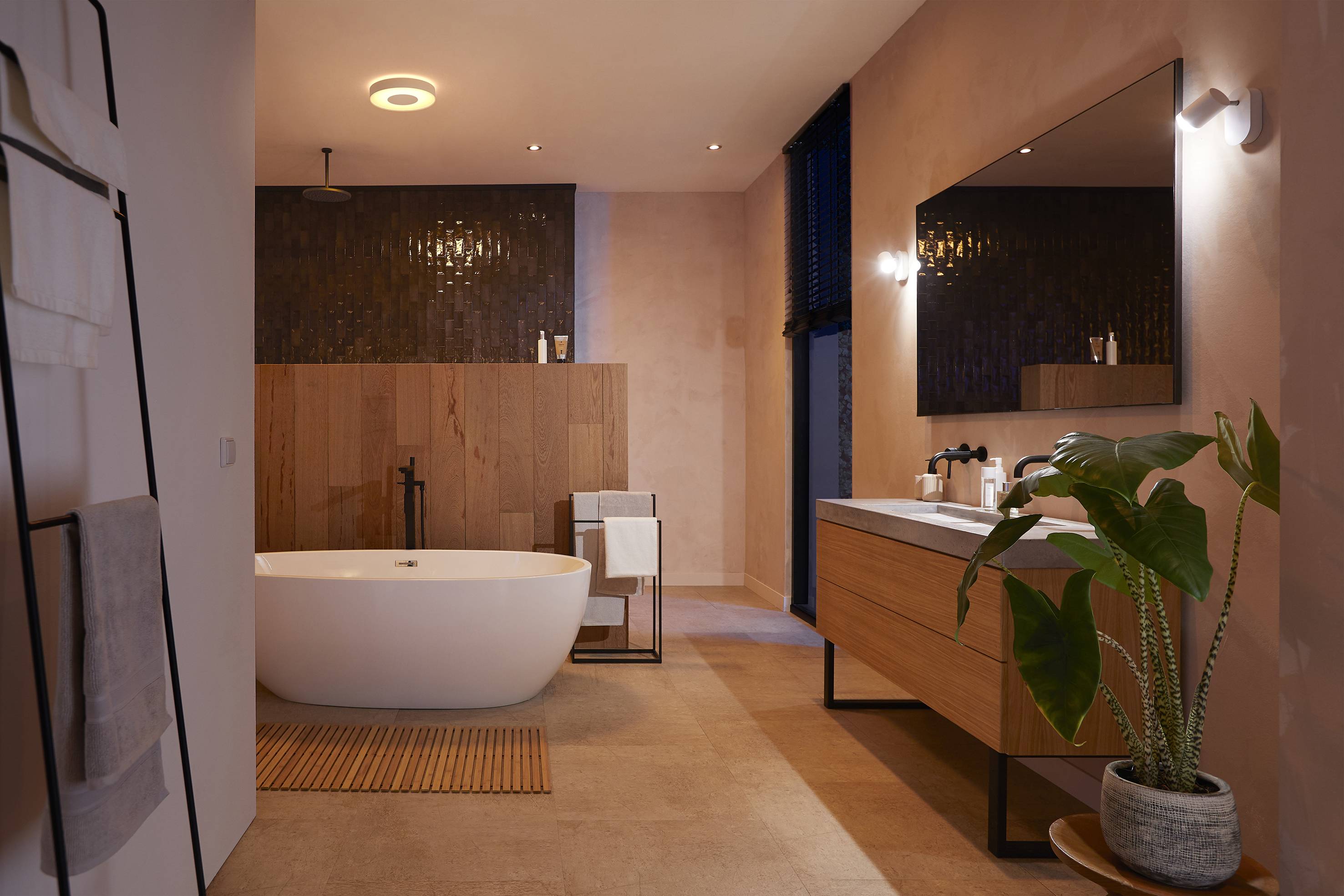 Modernes Badezimmer mit freistehender Badewanne, Holzverkleidung, großem Spiegel und Pflanze auf dem Boden, schummrige Beleuchtung.