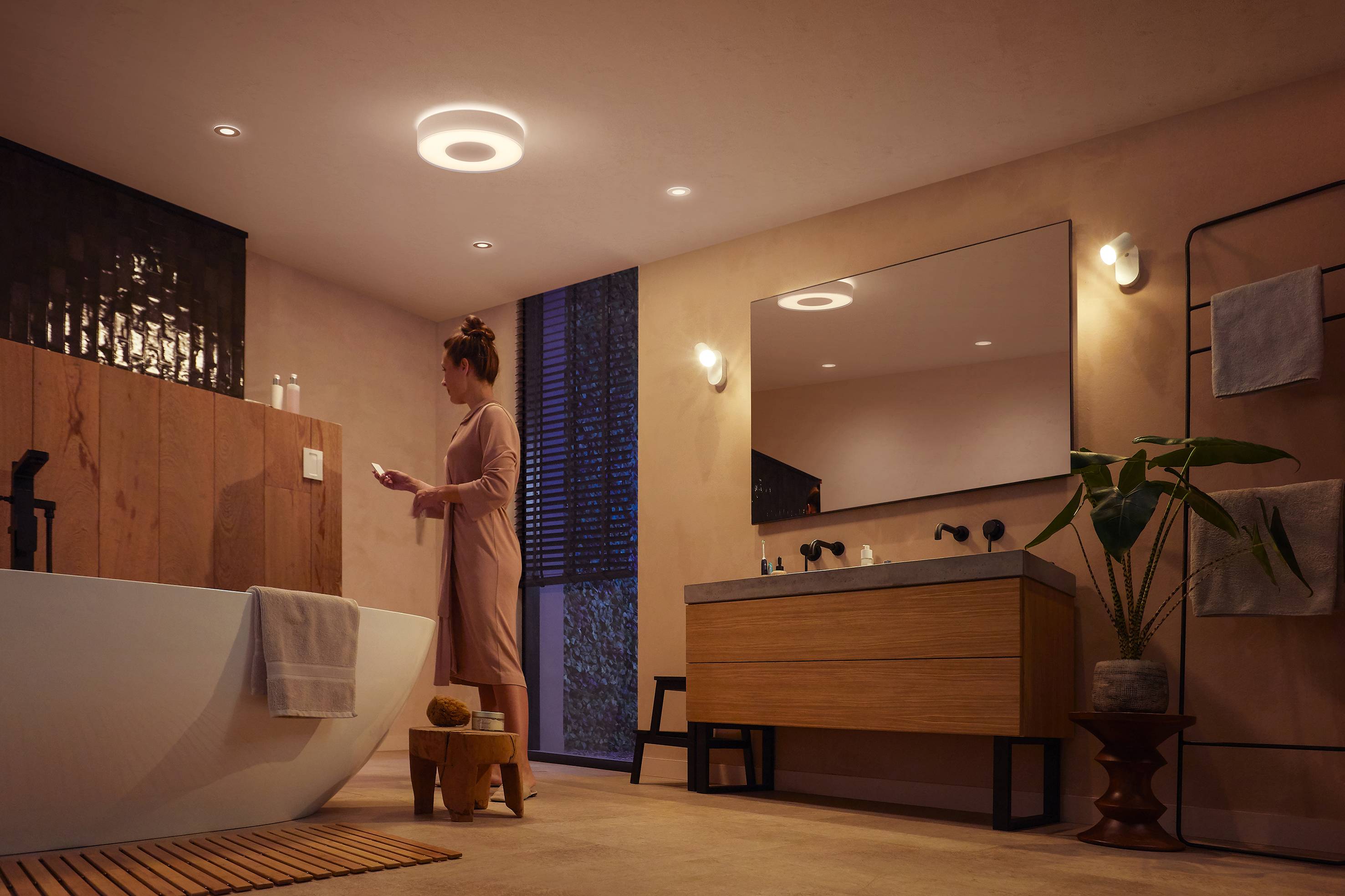 Eine Person steht in einem modernen Badezimmer mit Badewanne, Waschbecken und Pflanzen. Die Beleuchtung erzeugt eine warme Atmosphäre.