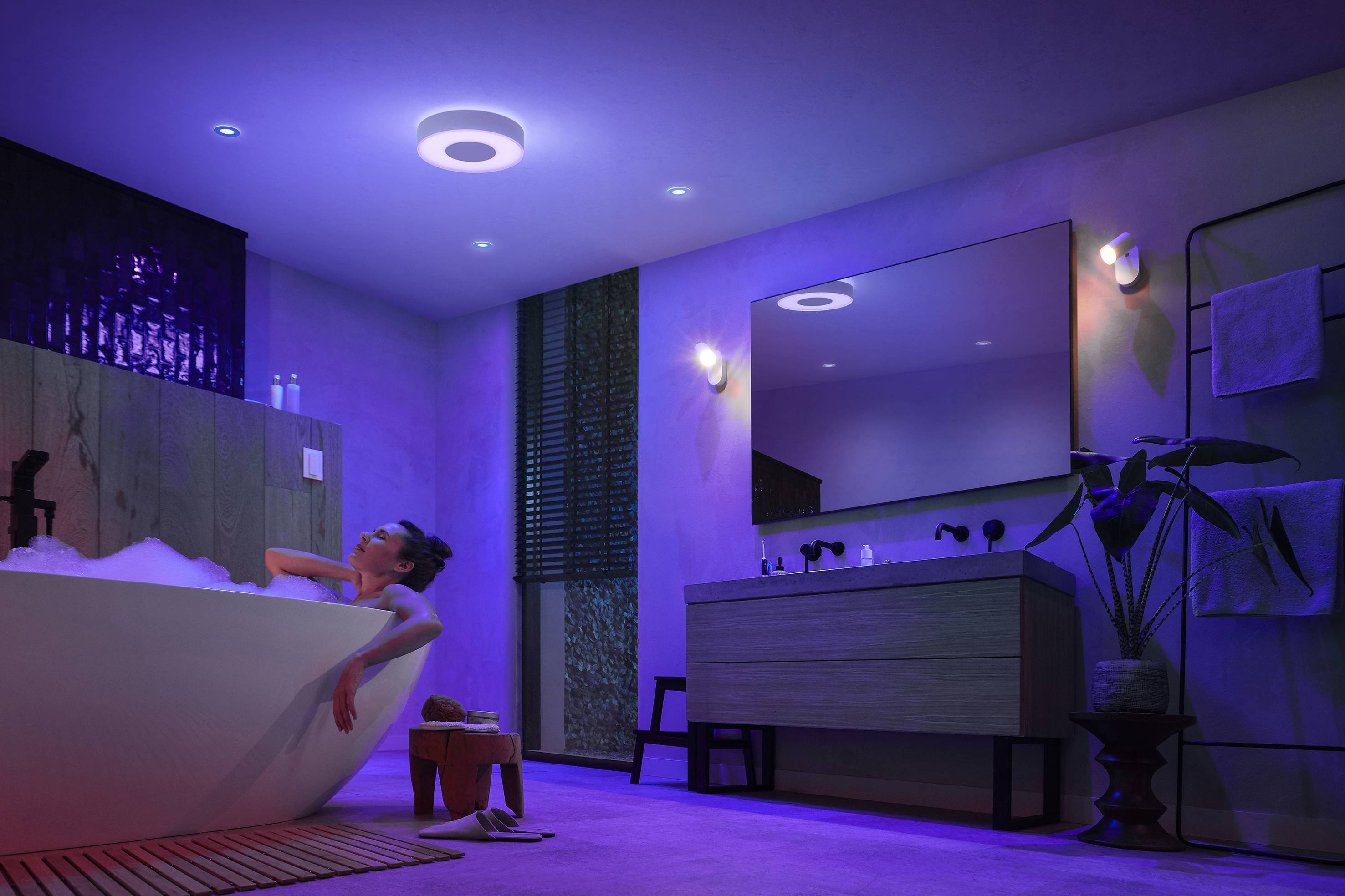 Eine Person entspannt in einer Badewanne in einem stimmungsvoll beleuchteten Badezimmer. Die Beleuchtung wechselt zwischen Blau und Violett.