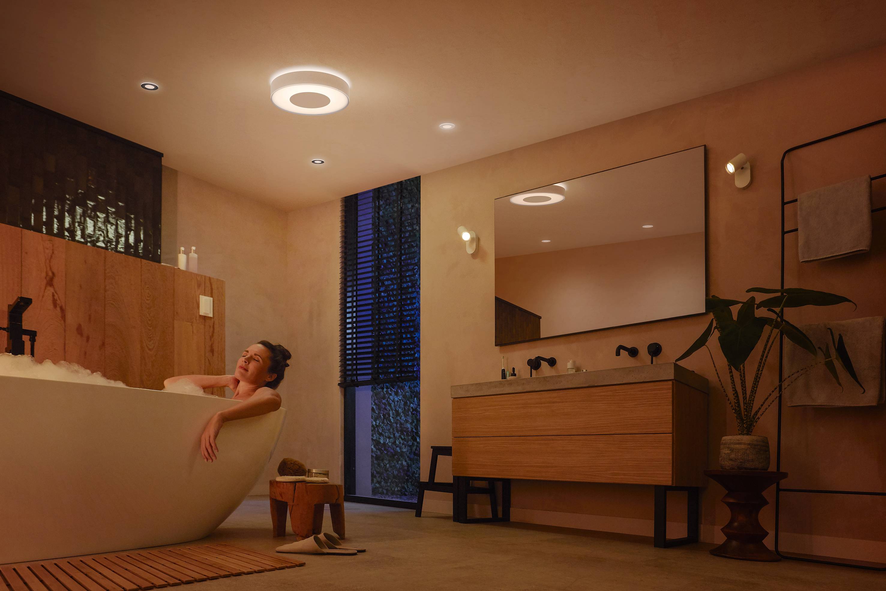 Eine Person entspannt sich in einem modernen Badezimmer mit dimmem Licht, liegend in einer weißen Badewanne, dekoriert mit Pflanzen.