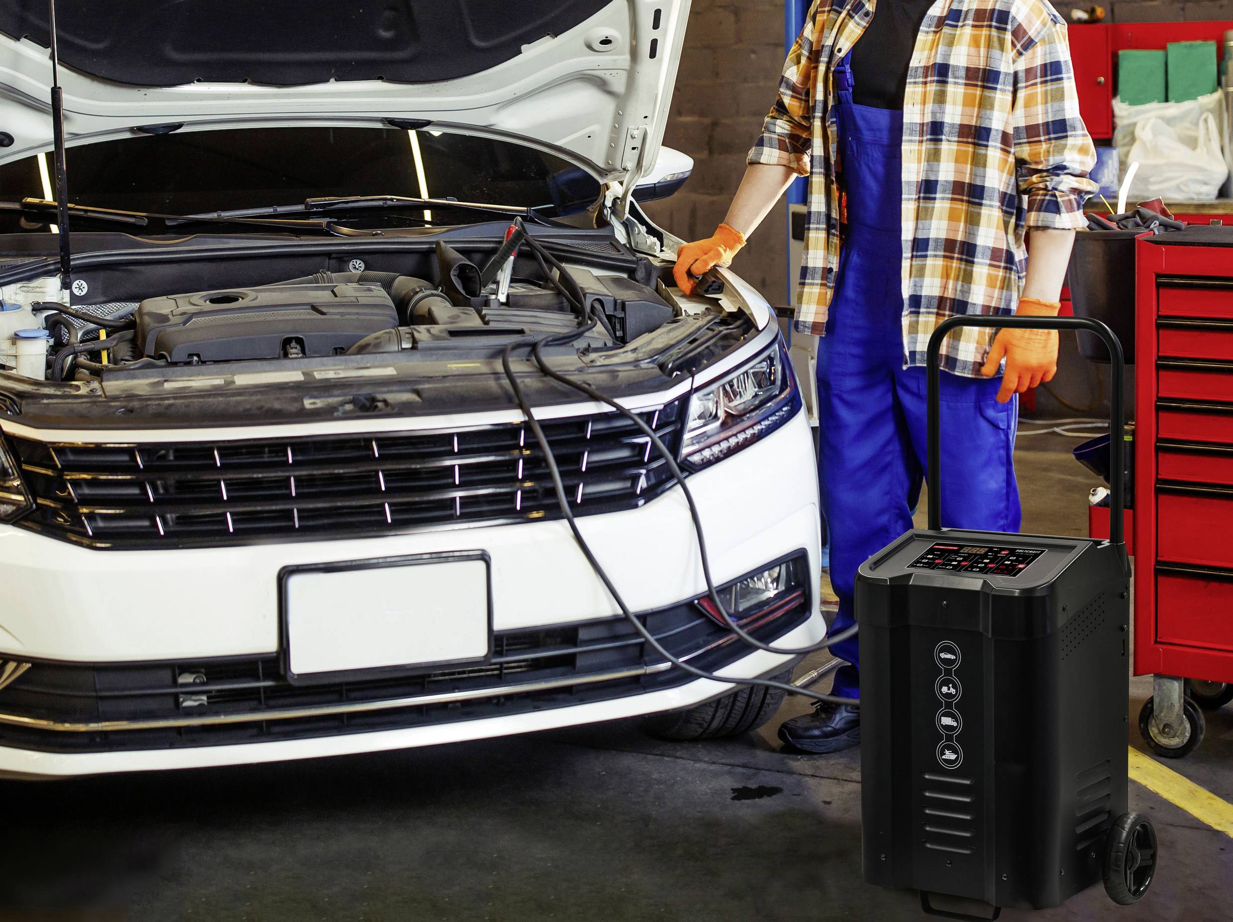 Ein Mechaniker steht neben einem Auto mit offener Motorhaube. Neben ihm befindet sich ein Batterieladegerät, verbunden mit der Autobatterie.