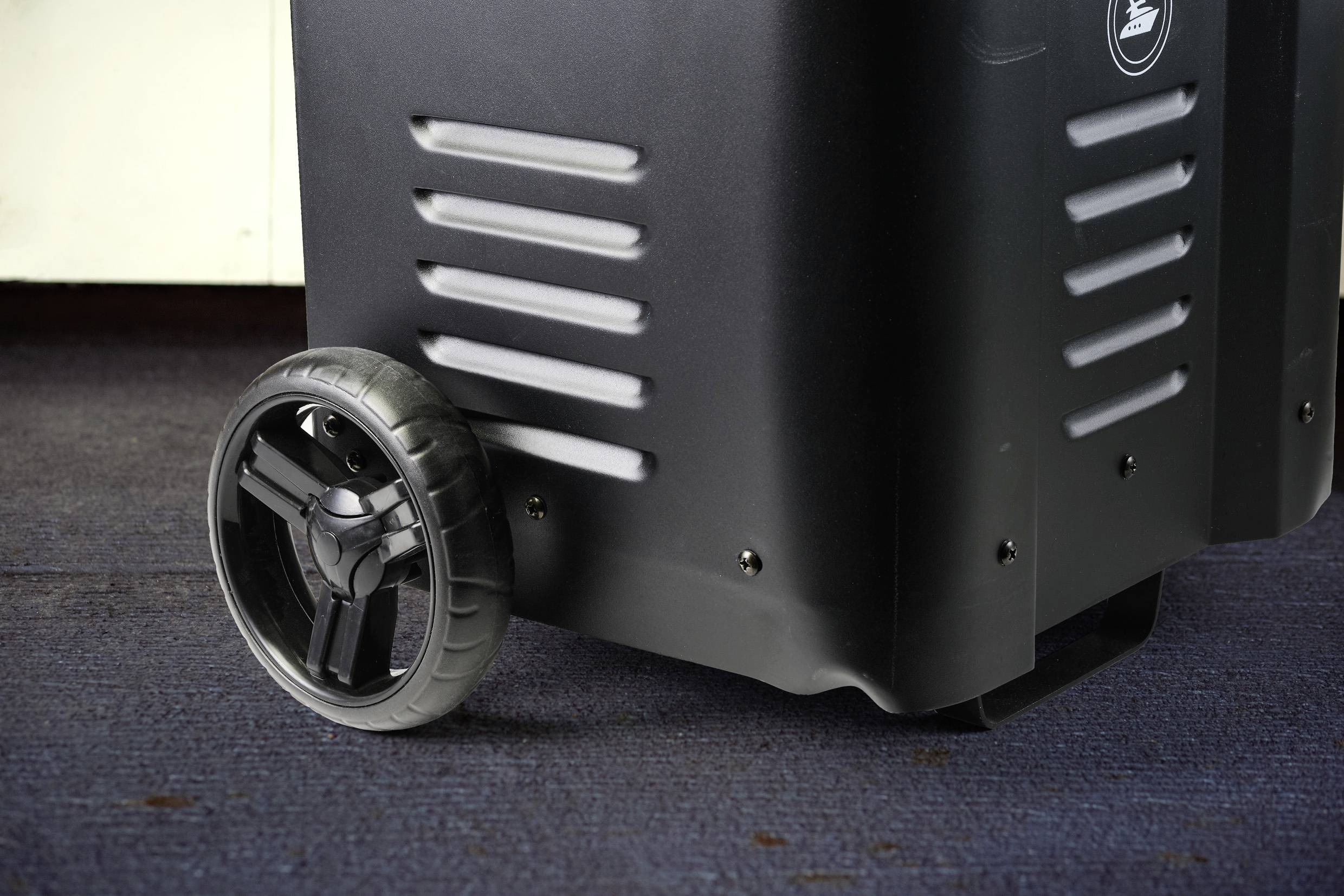 Schwarze Transportbox auf blauem Teppichboden mit einem großen Rad, geeignet für leichten Transport von Geräten oder Gepäck.