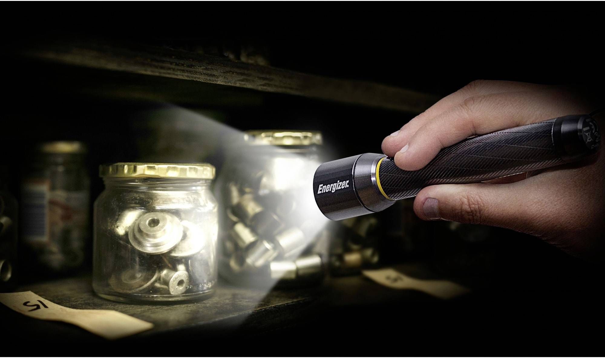 Eine Hand hält eine Taschenlampe und leuchtet auf Glasbehälter mit Metallteilen in einem dunklen Raum oder Lager.