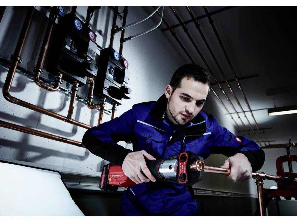 Eine Person in einer blauen Uniform verwendet ein rotes elektrisches Werkzeug, um an Kupferleitungen in einem industriellen Umfeld zu arbeiten, umgeben von Messgeräten und Rohren.