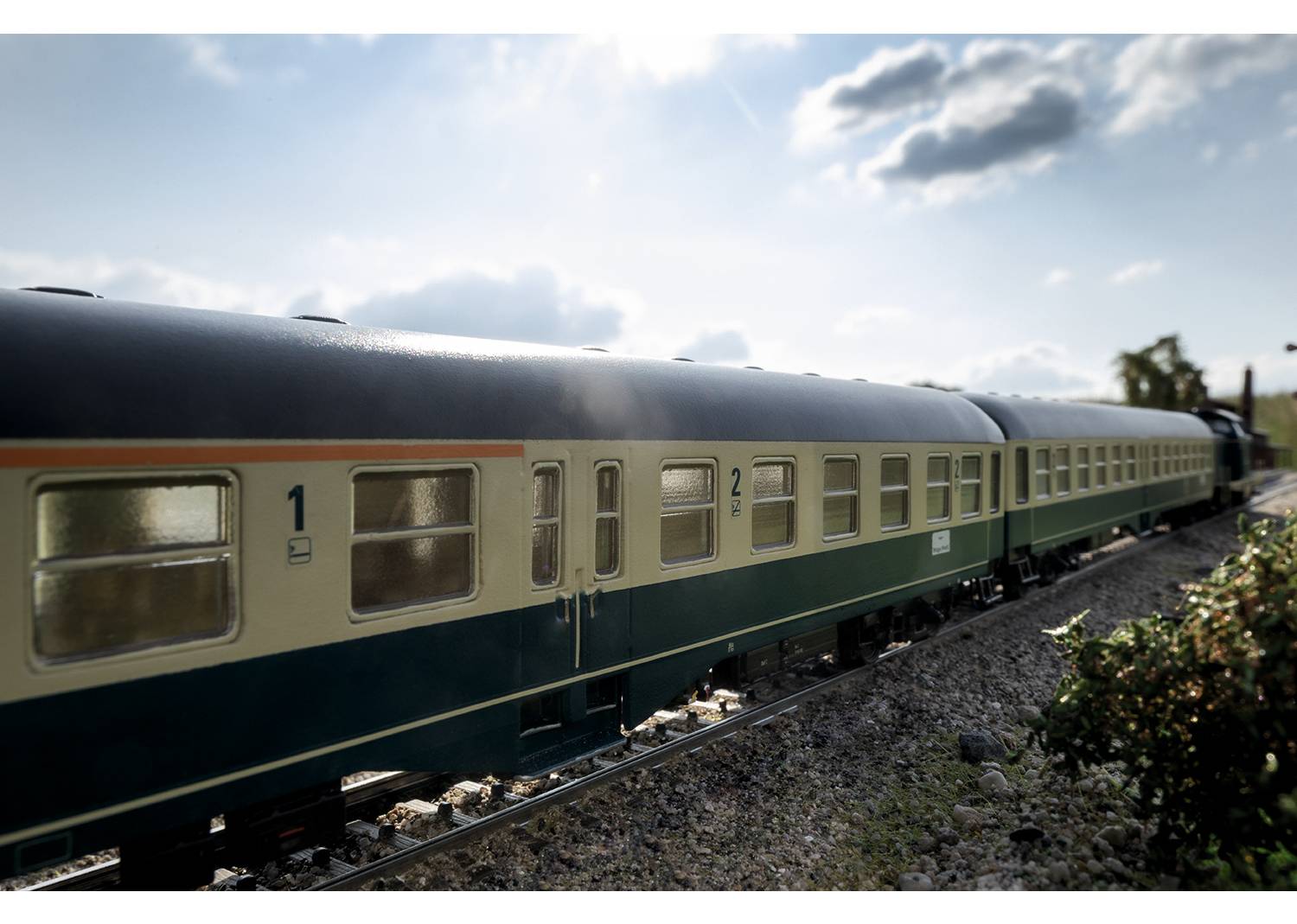 Ein Modellzug fährt auf einem Gleis durch eine Landschaft mit blauem Himmel und einigen Wolken. Die Waggons sind beige und blau gestrichen.