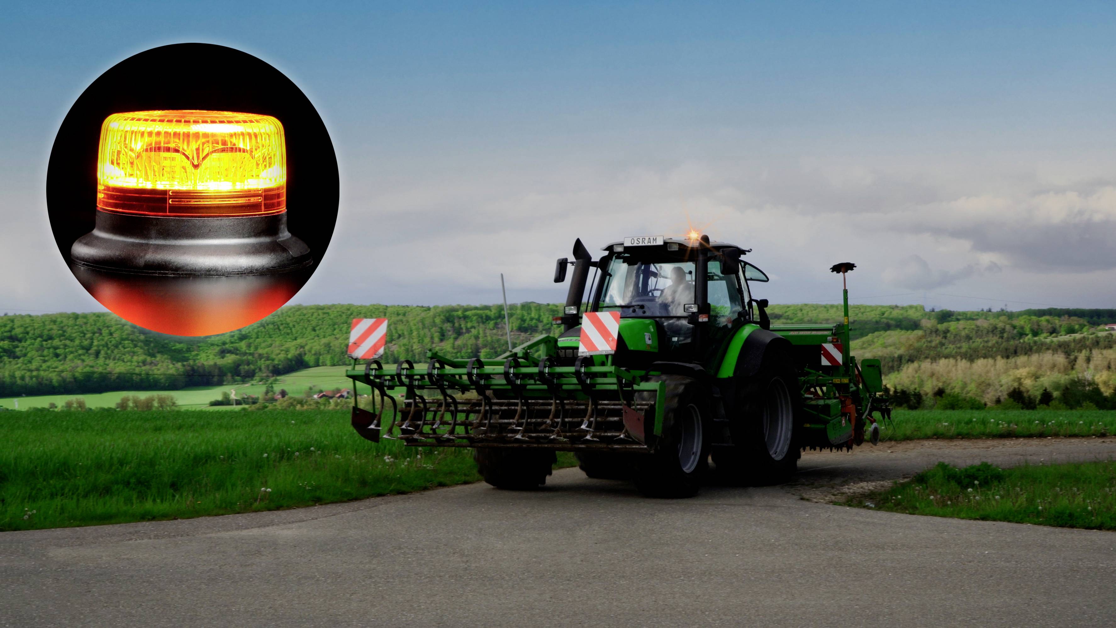 Ein Traktor auf einer Straße auf einem Feld mit einer Nahaufnahme eines gelben Warnlichts. Wälder und Himmel im Hintergrund.