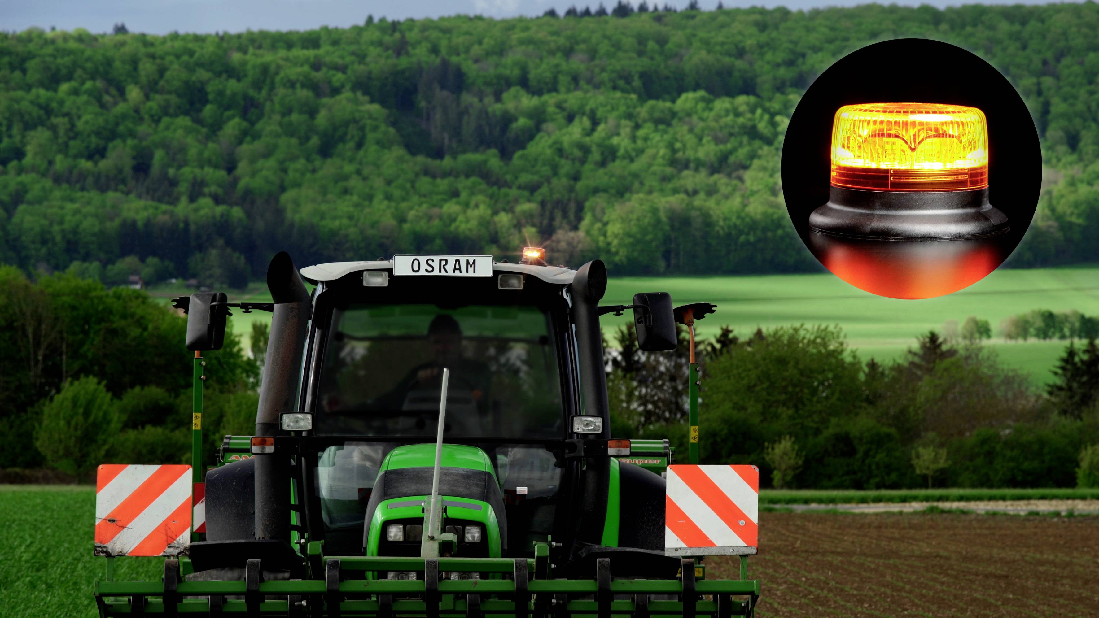 Ein Traktor auf einem Feld vor einem Waldhintergrund. Im oberen Teil ist ein oranges Warnlicht vergrößert dargestellt.