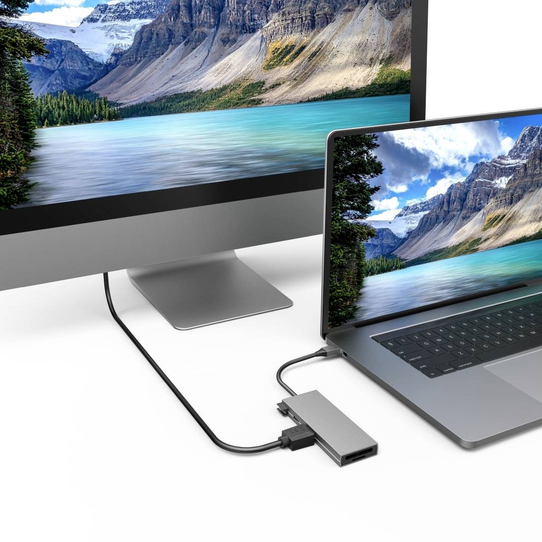 Ein Laptop ist über einen Adapter mit einem Monitor verbunden, beide zeigen eine identische Landschaft mit Bergen und einem See.