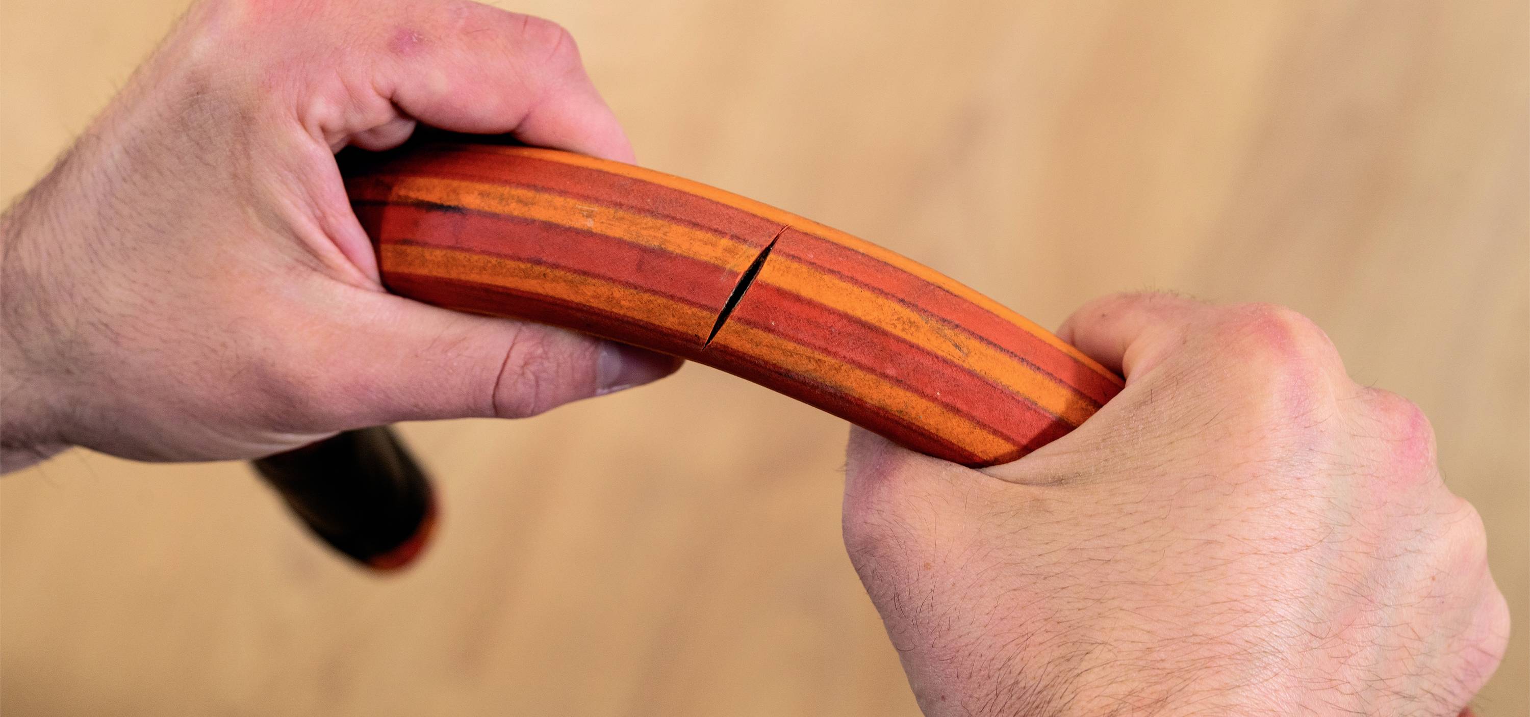 Eine Hand bricht ein ovales, mehrfarbiges Holzobjekt. Die Szene zeigt Kraft oder Anstrengung. Hintergrund ist unscharf.