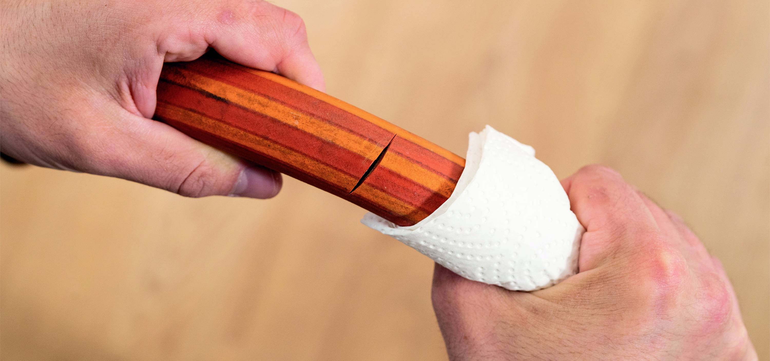 Zwei Hände greifen ein rotes Holzstab, der eine weiße Papierschablone aufweist, auf einem hellbraunen Hintergrund.