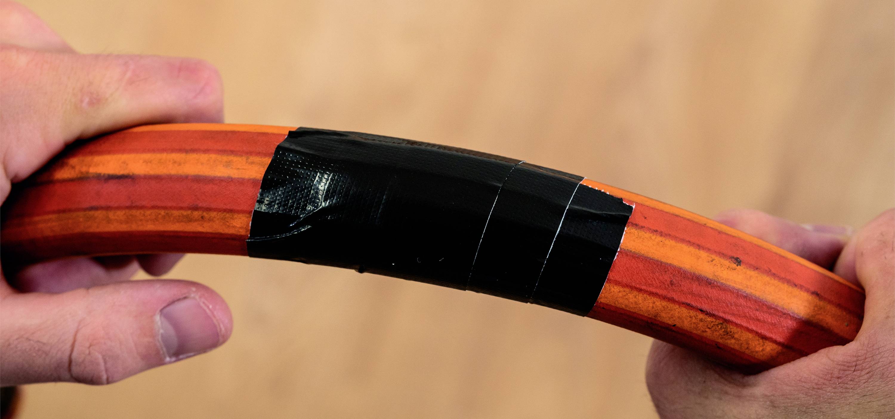 Zwei Hände halten ein mit schwarzem Klebeband umwickeltes gebogenes Rohr. Das Rohr ist orange mit roten Streifen.