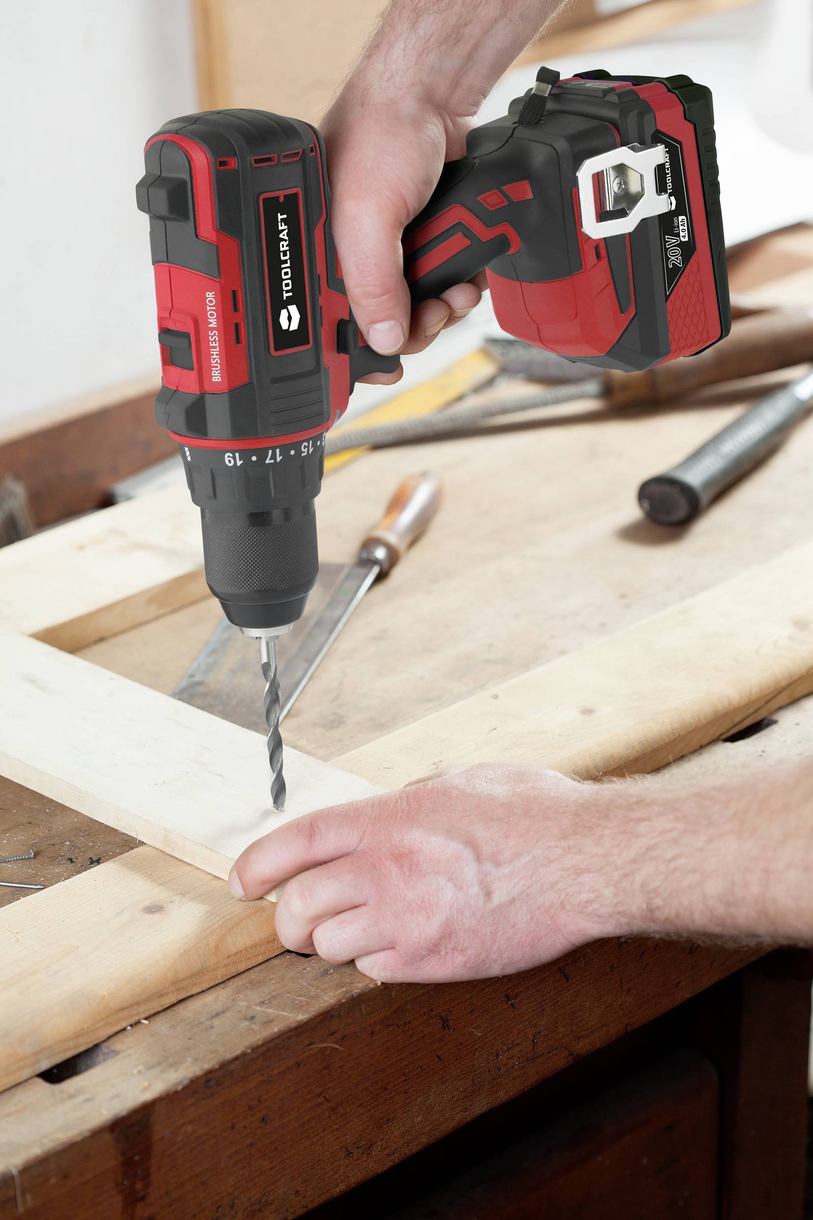 Eine Person bohrt mit einem roten Akkuschrauber ein Loch in ein Holzbrett auf einer Werkbank. Werkzeuge liegen daneben.