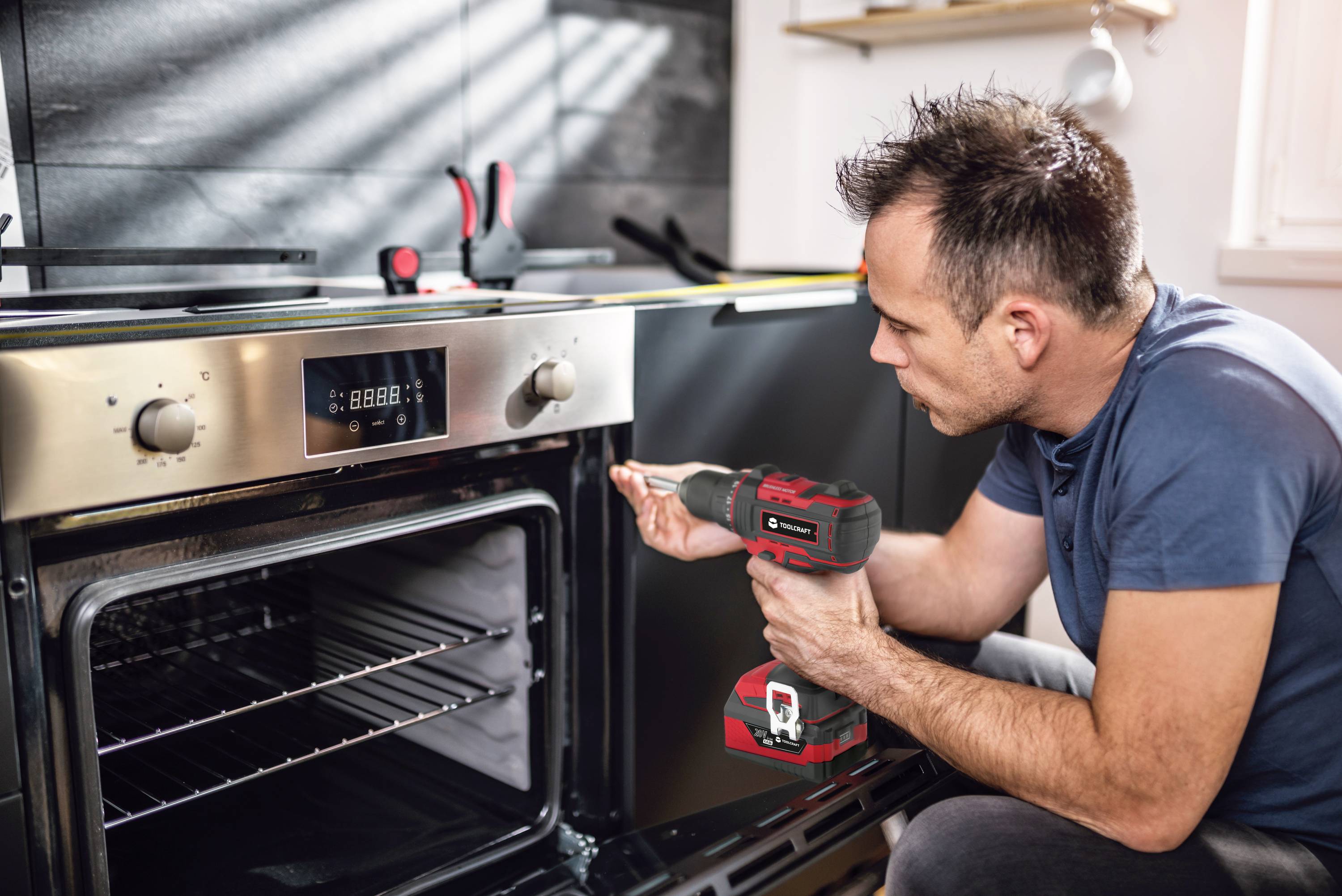 Ein Mann repariert einen Ofen mit einem Akkubohrer in einer modernen Küche. Werkzeuge liegen auf der Arbeitsfläche.