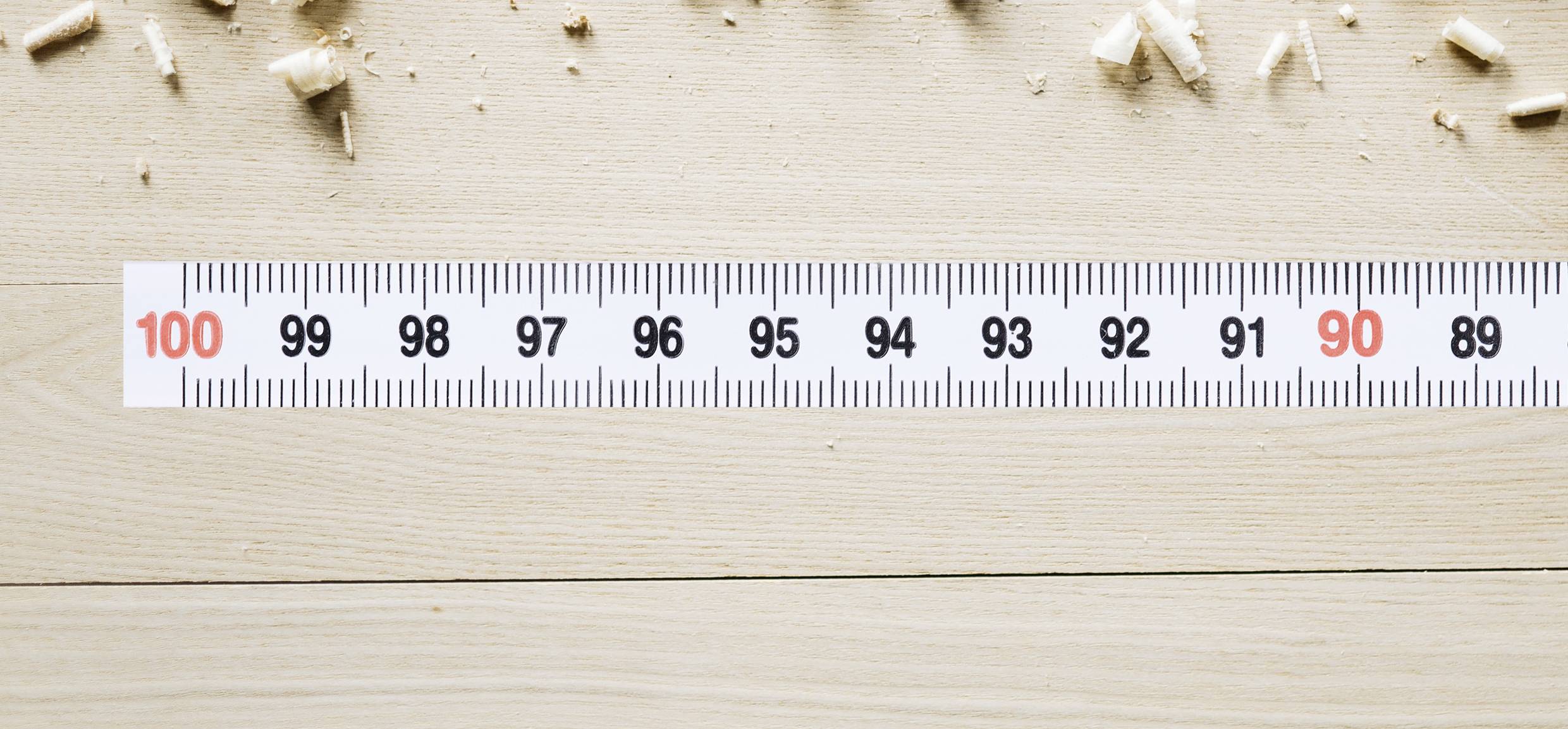 Ein Maßband liegt flach auf einem Holzboden, mit Markierungen von 89 bis 100 sichtbar. Holzspäne sind um das Maßband verstreut.
