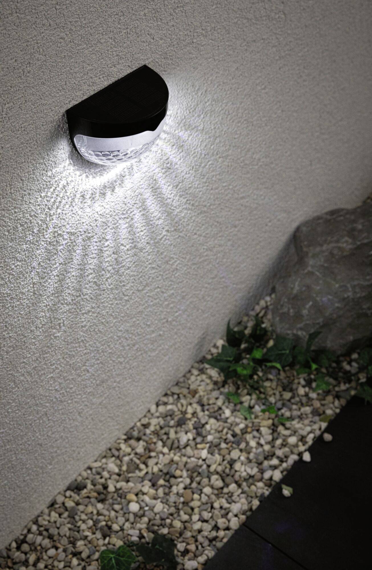 Wandleuchte beleuchtet eine Steinwand und verstreut Lichtstrahlen. Darunter sind Kieselsteine und ein großer Stein sichtbar.