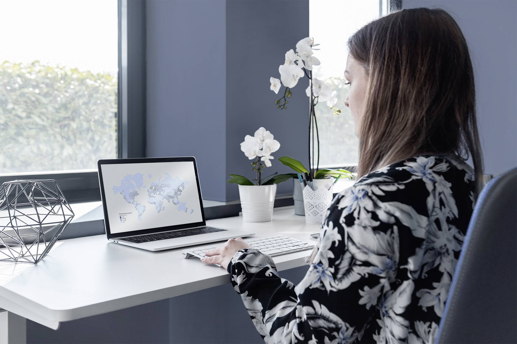 Frau sitzt an einem Tisch und arbeitet am Laptop. Der Bildschirm zeigt eine Weltkarte. Orchideen und geometrische Dekorationen sind sichtbar.