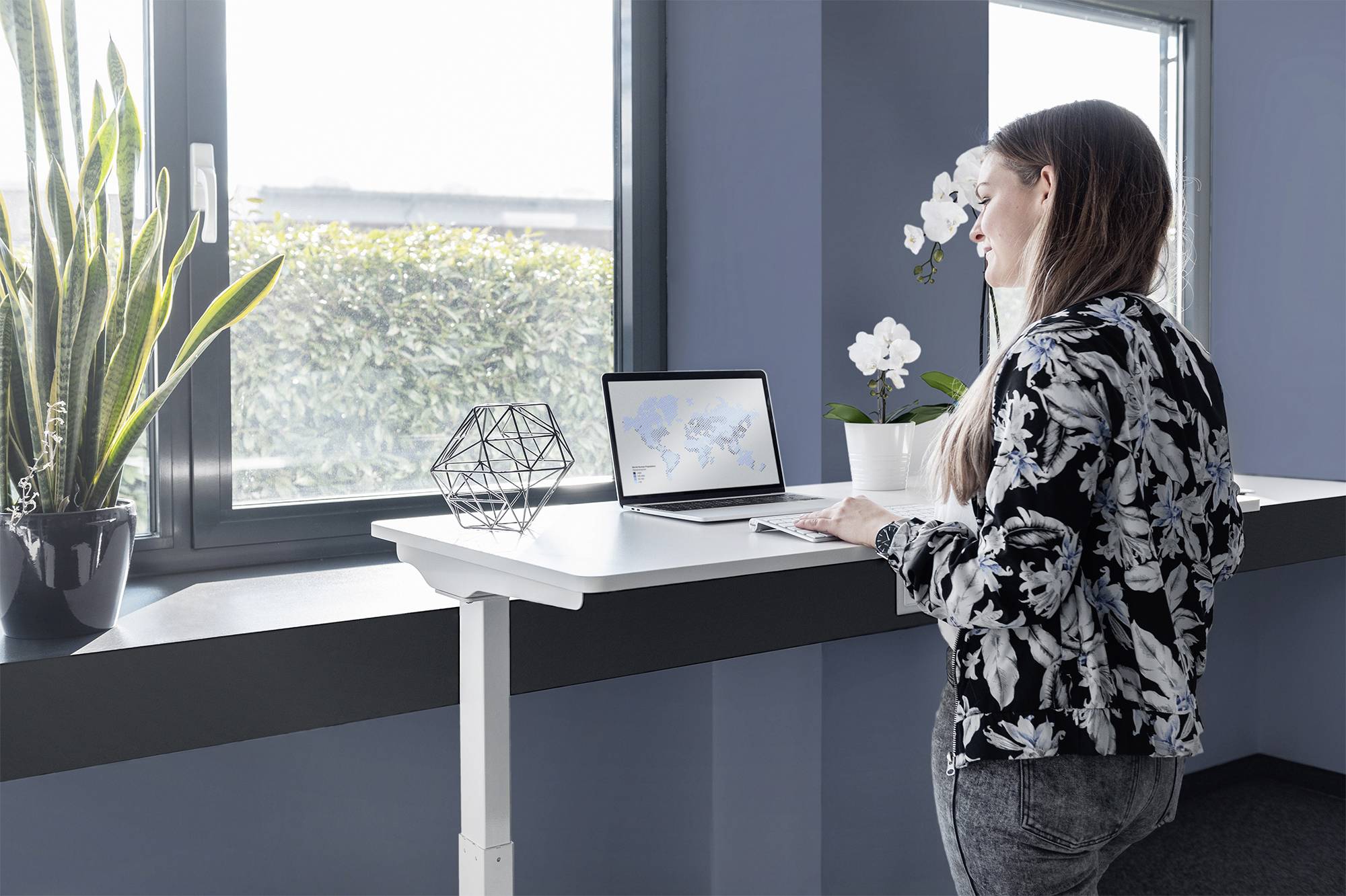 Eine Frau steht an einem höhenverstellbaren Schreibtisch und arbeitet am Laptop. Im Hintergrund sind Pflanzen und ein Fenster zu sehen.