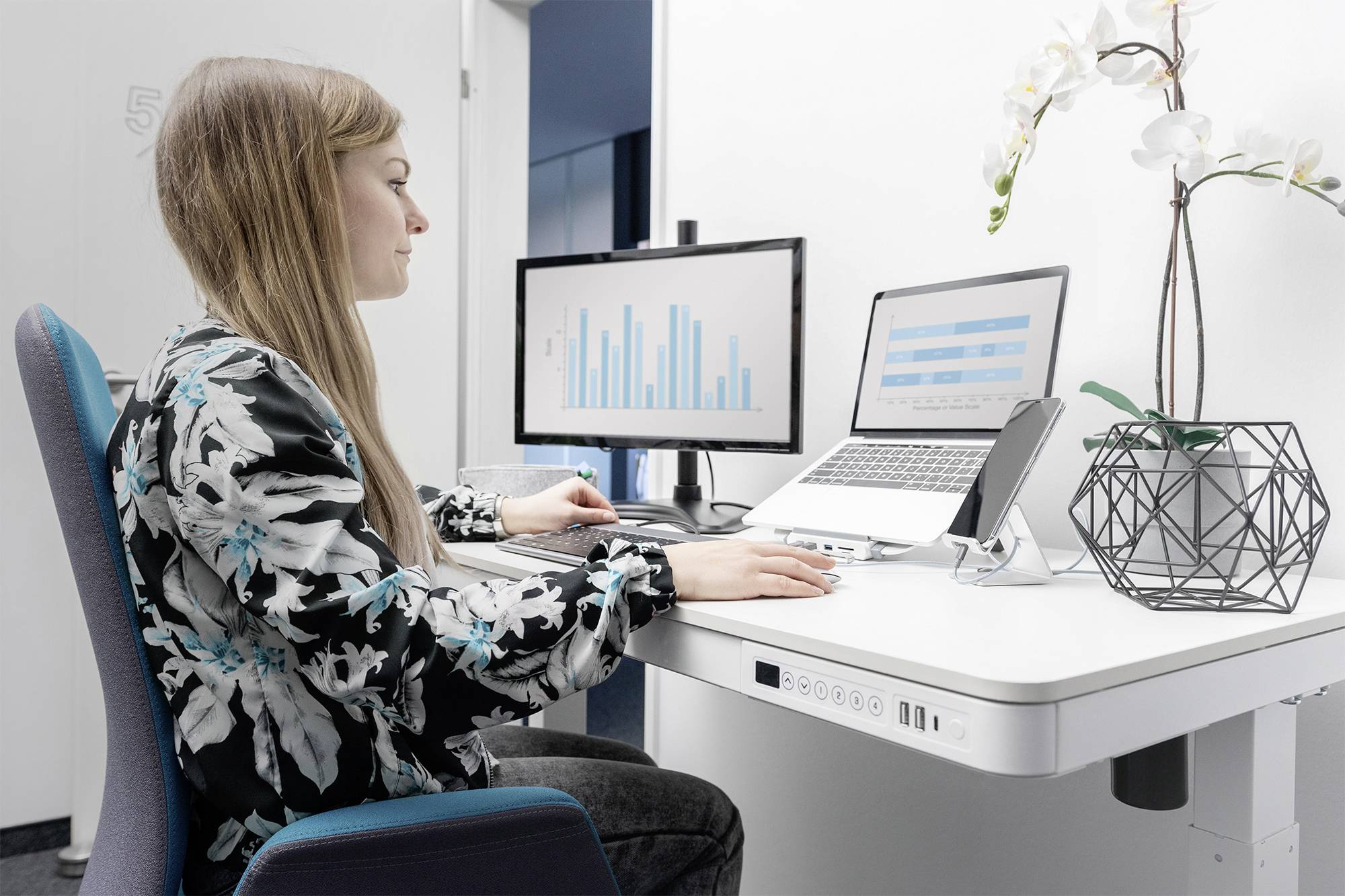 Eine Frau sitzt an einem Schreibtisch und arbeitet am Laptop. Auf dem Monitor sind Balkendiagramme sichtbar. Daneben steht eine Orchidee.