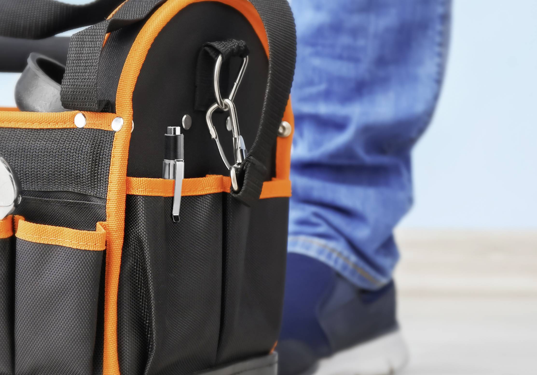 Eine schwarze Werkzeugtasche mit orangefarbenen Nähten steht auf einem Boden. Eine Person in blauen Jeans und Sneakers steht daneben.