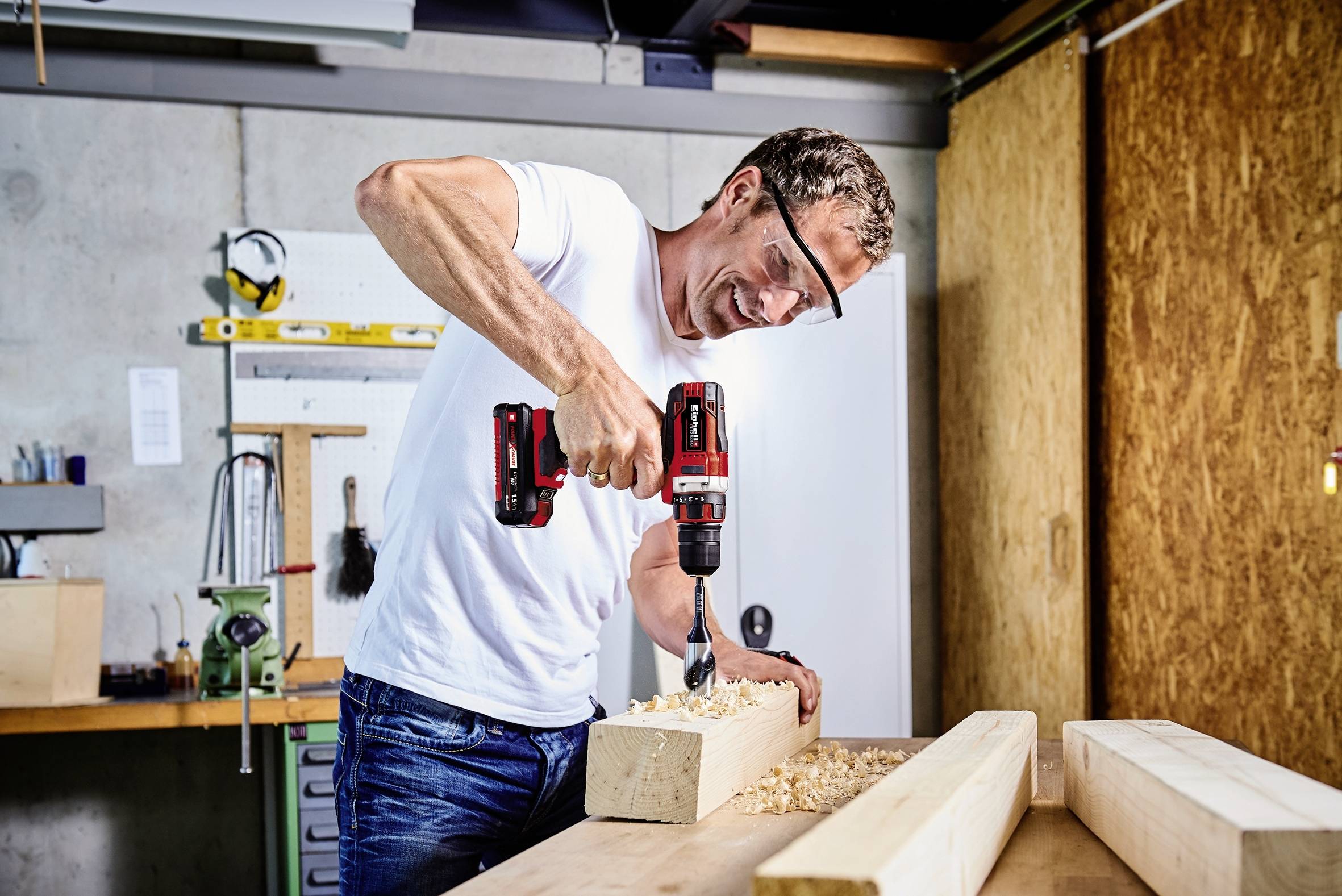 Ein Mann in Werkstatt bohrt lachend ein Loch in einen Holzbalken. Werkzeuge und Holzarbeiten im Hintergrund.