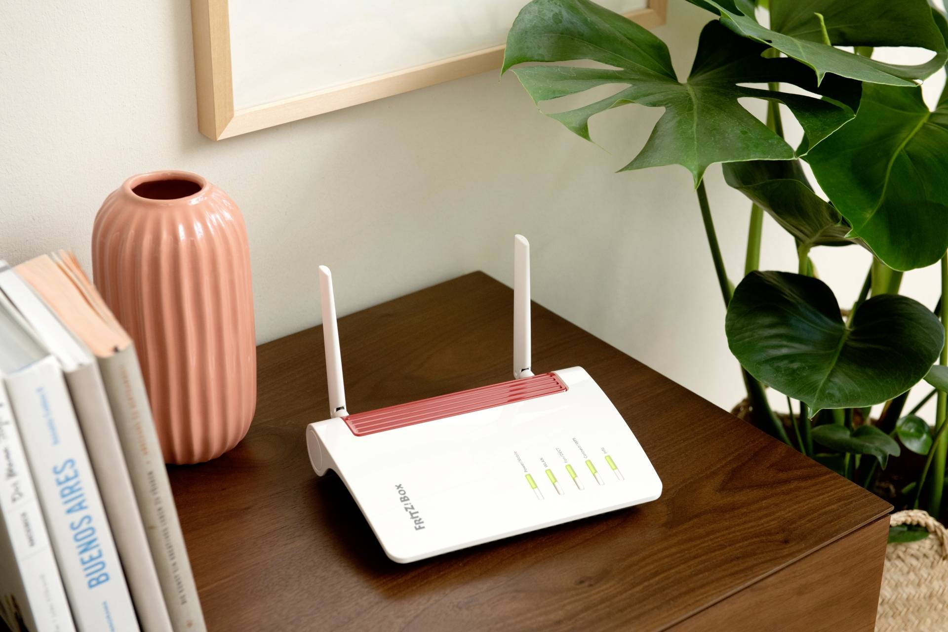 Ein weißer Router mit Antennen und vier leuchtenden LEDs steht auf einem Holztisch neben Büchern und einer Pflanze.