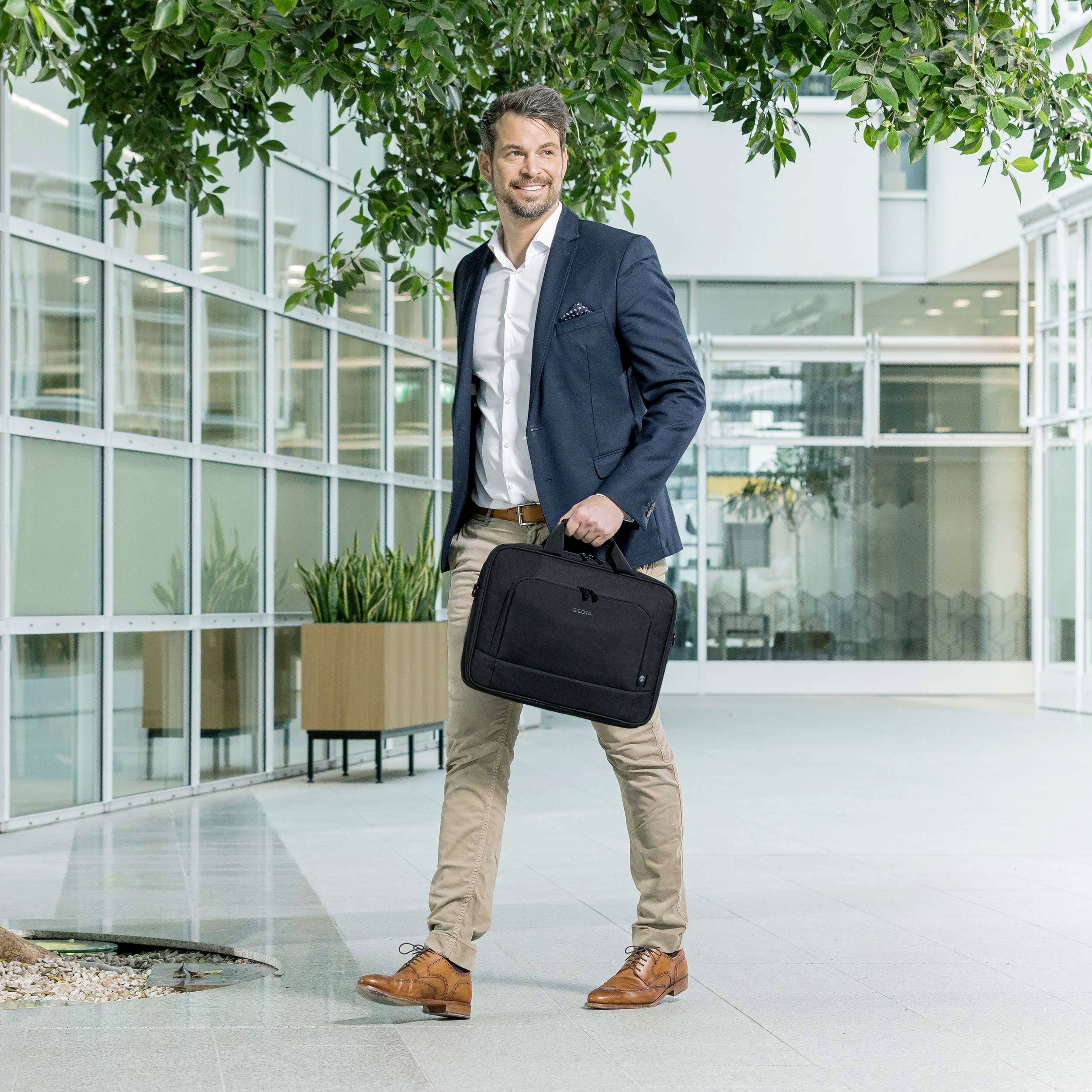 Mann im Business-Outfit geht lächelnd durch modernes Bürogebäude, trägt eine schwarze Tasche in der Hand.