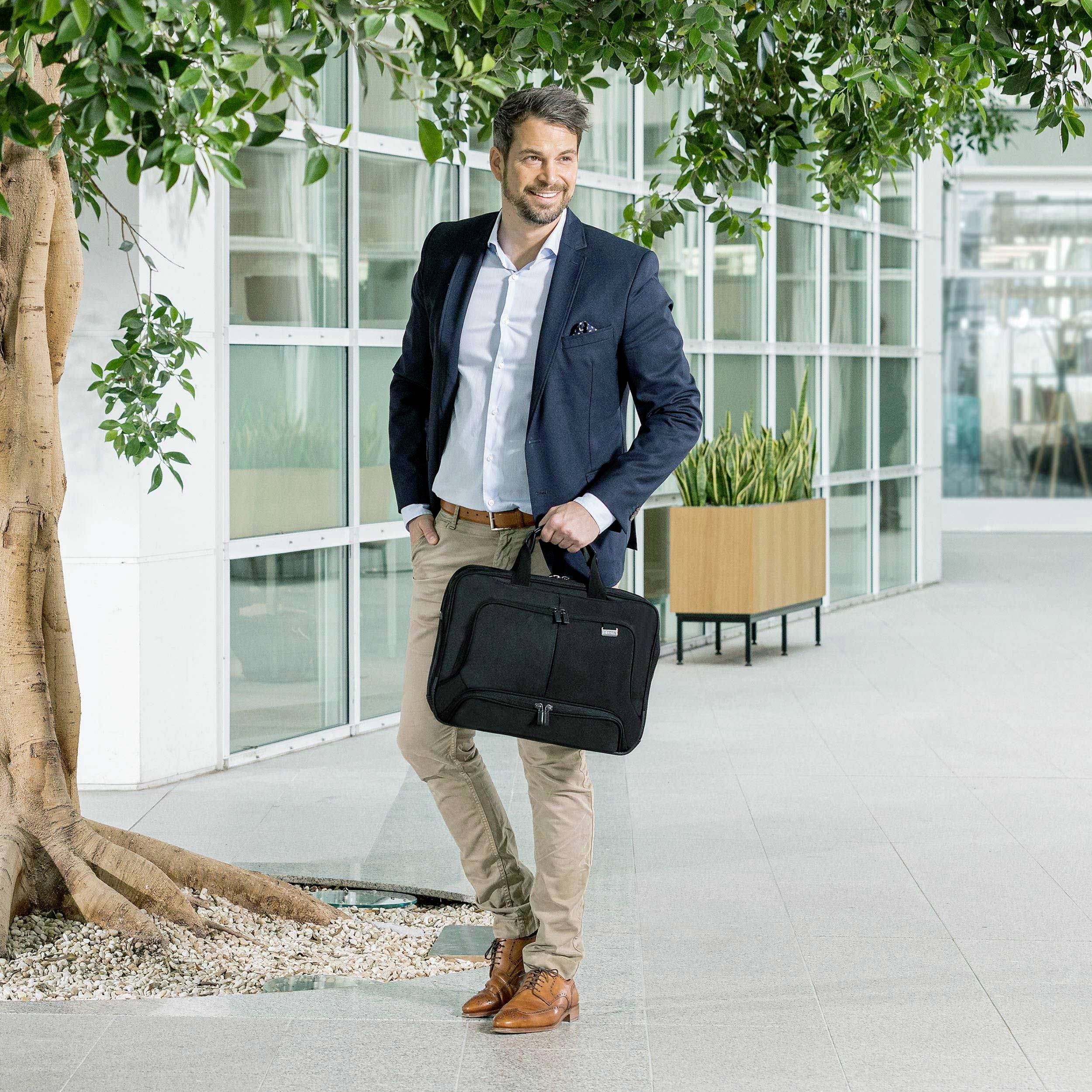 Ein Mann in Business-Kleidung geht lächelnd in einem modernen Bürogebäude entlang. Er trägt eine schwarze Laptoptasche in der rechten Hand.