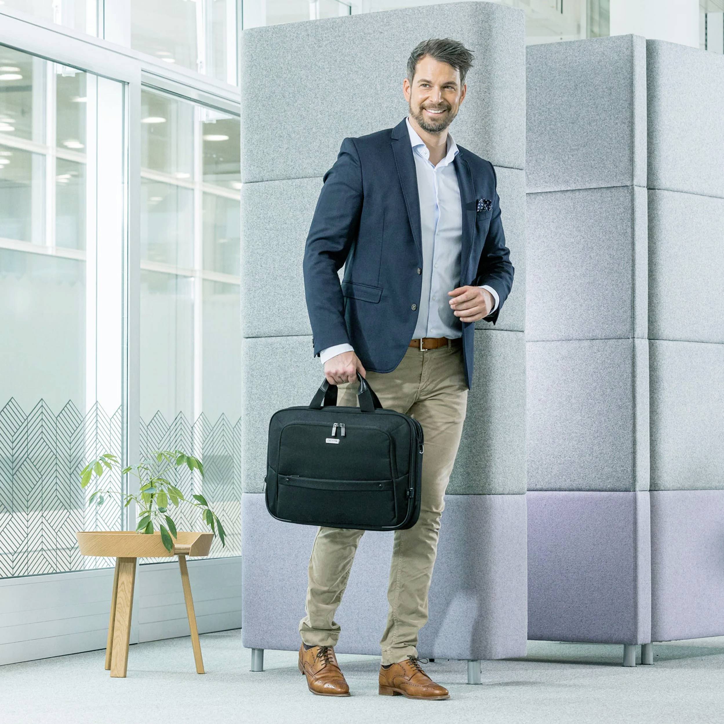 Ein Mann im Anzug steht lächelnd neben einer grauen Wand, hält eine schwarze Tasche. Im Hintergrund sind große Fenster und ein Tisch mit einer Pflanze.