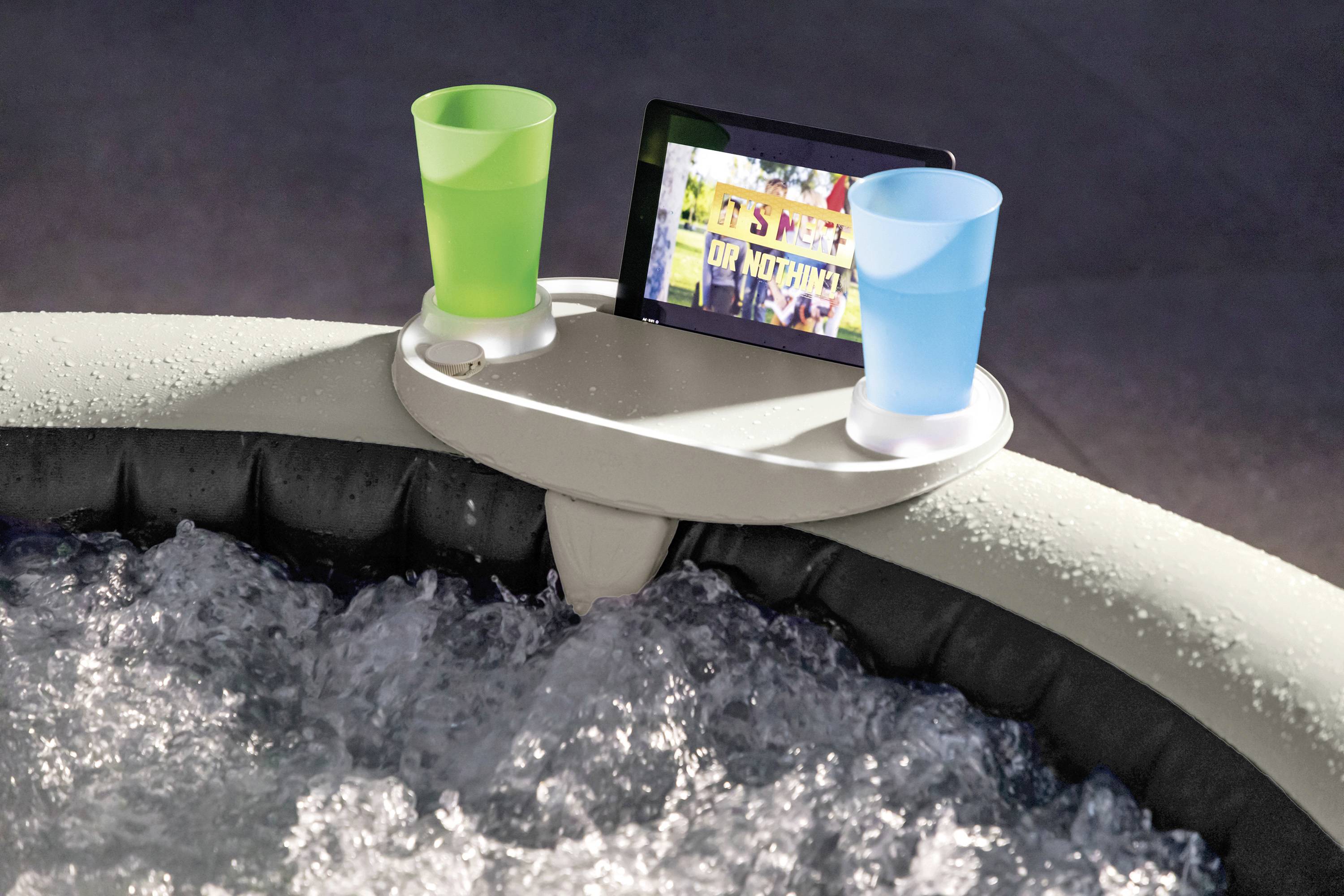 Zwei bunte Becher und ein Tablet stehen auf einer Halterung am Rand eines Whirlpools mit sprudelndem Wasser.