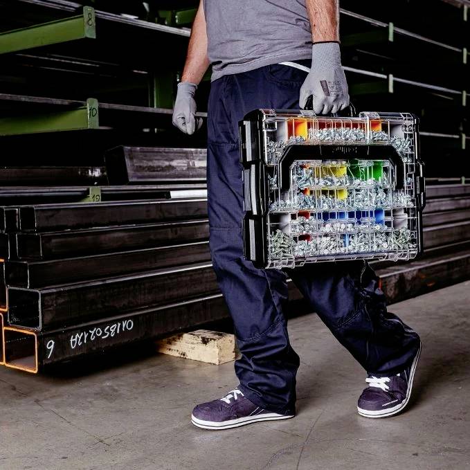 Eine Person mit Handschuhen hält eine Kunststoff-Werkzeugbox mit farbigen Fächern und steht neben Metallregalen.