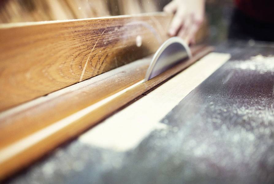Eine Hand führt ein Holzbrett gegen ein rotierendes Sägeblatt auf einem Tisch, um es zu schneiden. Sägemehl liegt auf der Oberfläche.