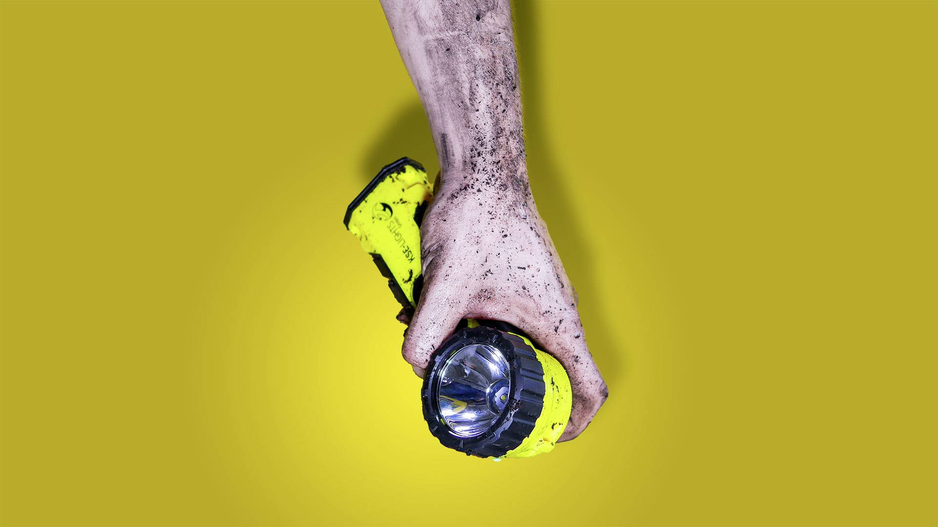 Eine schlammige Hand hält eine gelbe Taschenlampe vor einem gelben Hintergrund.