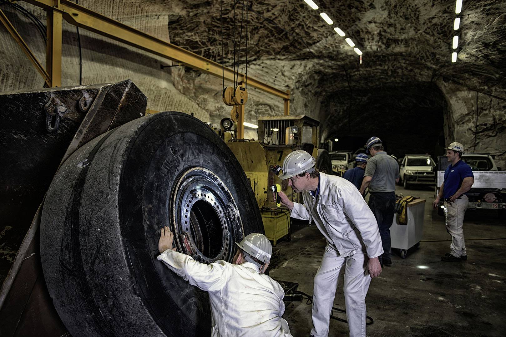 Arbeiter in einem Bergwerk reparieren ein riesiges Maschinenrad. Im Hintergrund befindet sich ein dunkler Stollen.