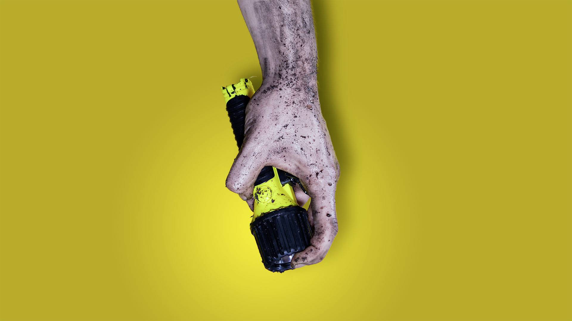 Eine Hand mit Erde bedeckt hält eine gelbe und schwarze Taschenlampe vor einem gelben Hintergrund.