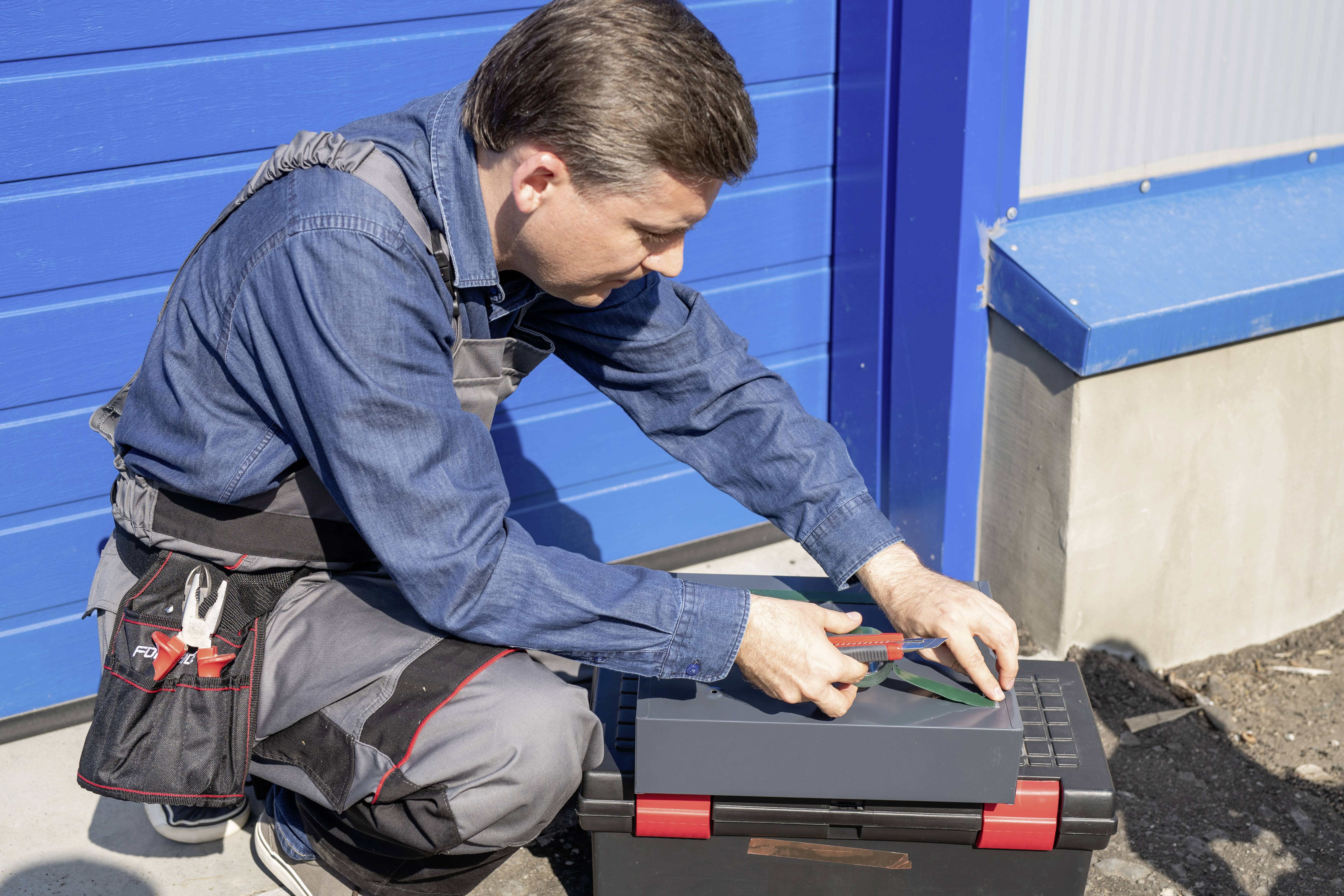 Ein Mann in Arbeitskleidung sitzt auf einem Werkzeugkasten vor einem blauen Garagentor und schneidet etwas mit einem Cuttermesser.