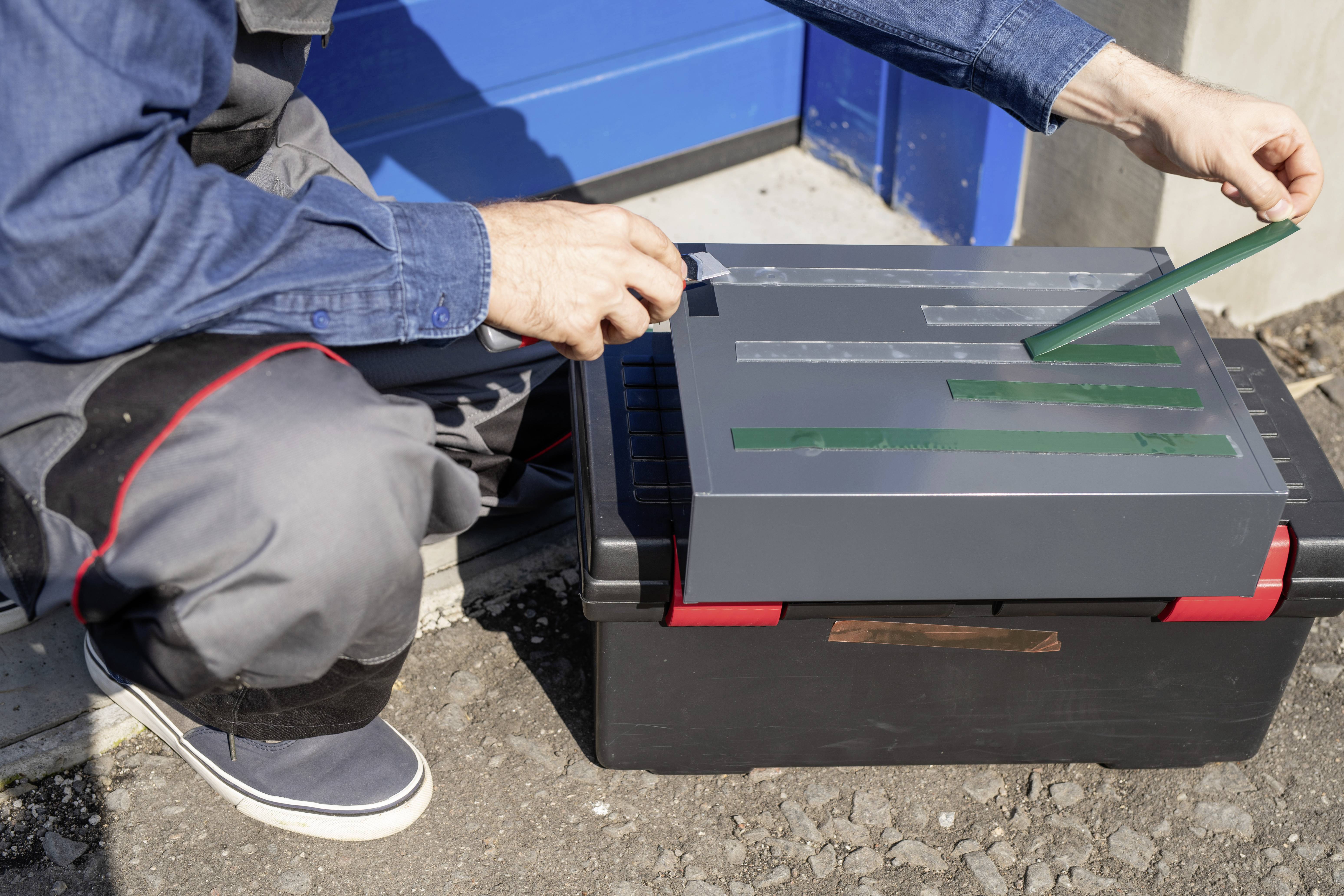 Eine Person klebt grüne Streifen auf eine graue Box, die auf einer schwarzen Kiste steht. Hintergrund zeigt eine blaue Tür.