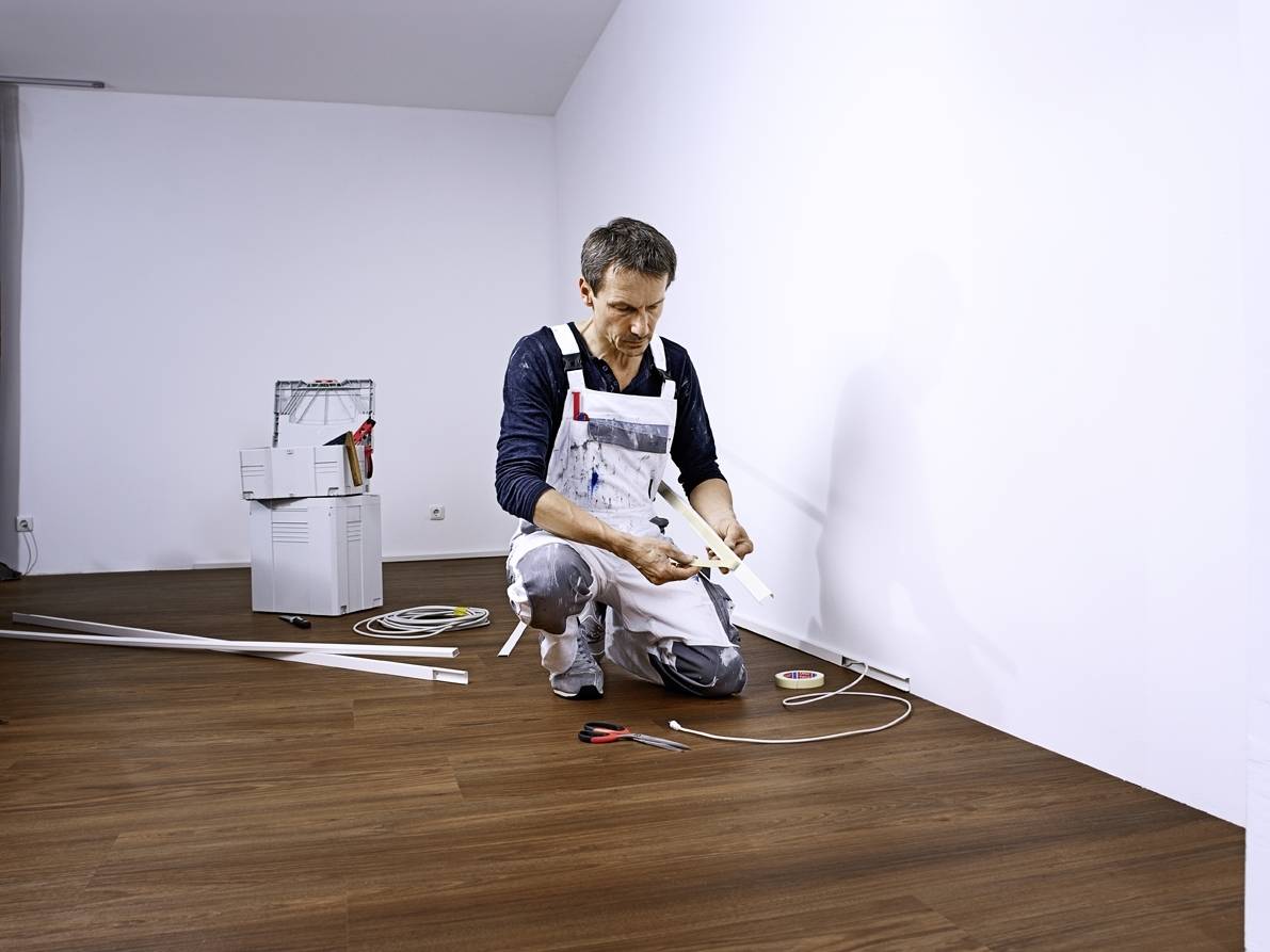 Ein Handwerker kniet auf einem Holzboden und arbeitet mit Kabeln und Werkzeugen. Im Hintergrund steht ein Kopierer.