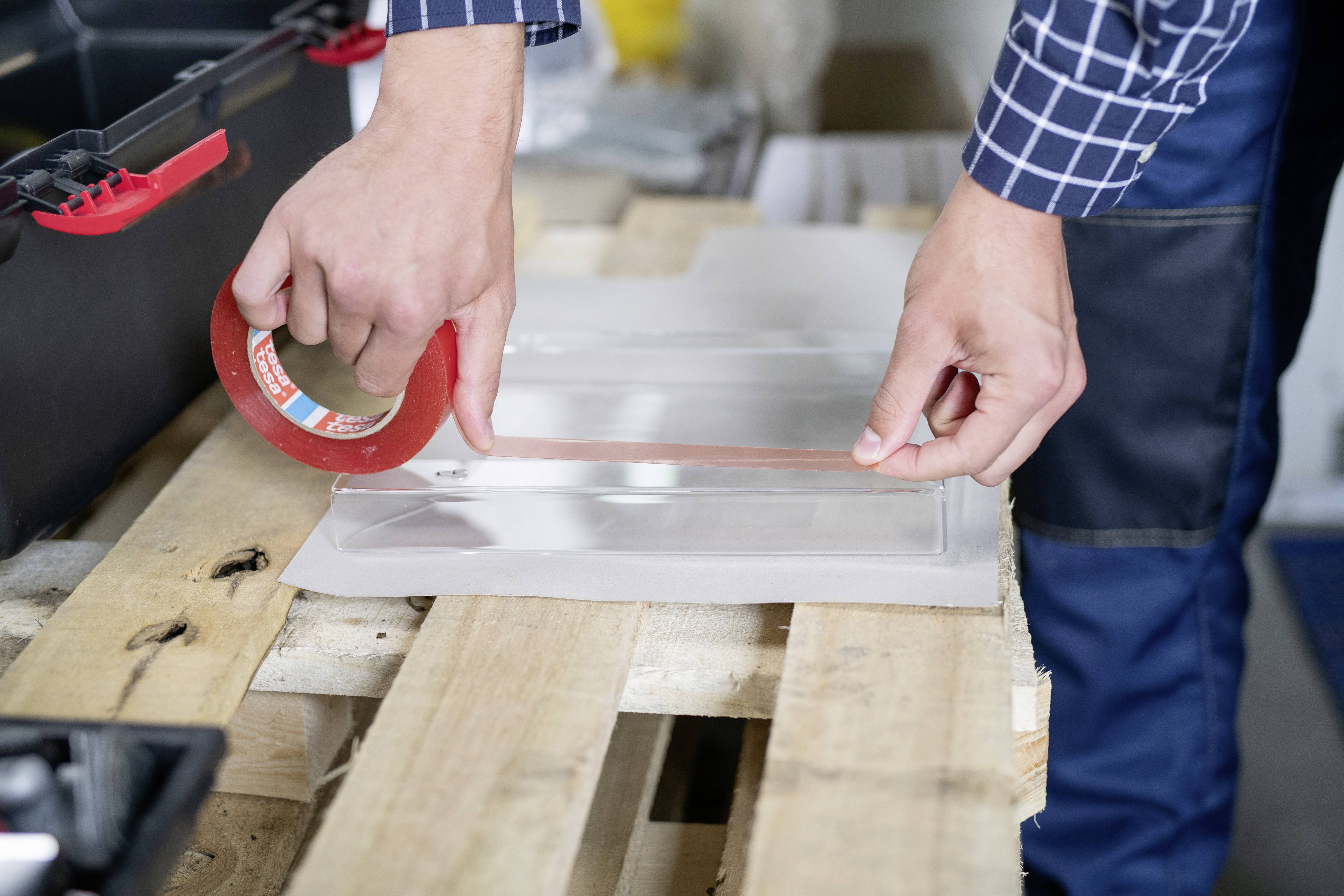 Person klebt rotes Klebeband auf ein Stück Glas oder Acryl, das auf einem Holzboden liegt. Neben der Person steht ein geöffneter Werkzeugkasten.