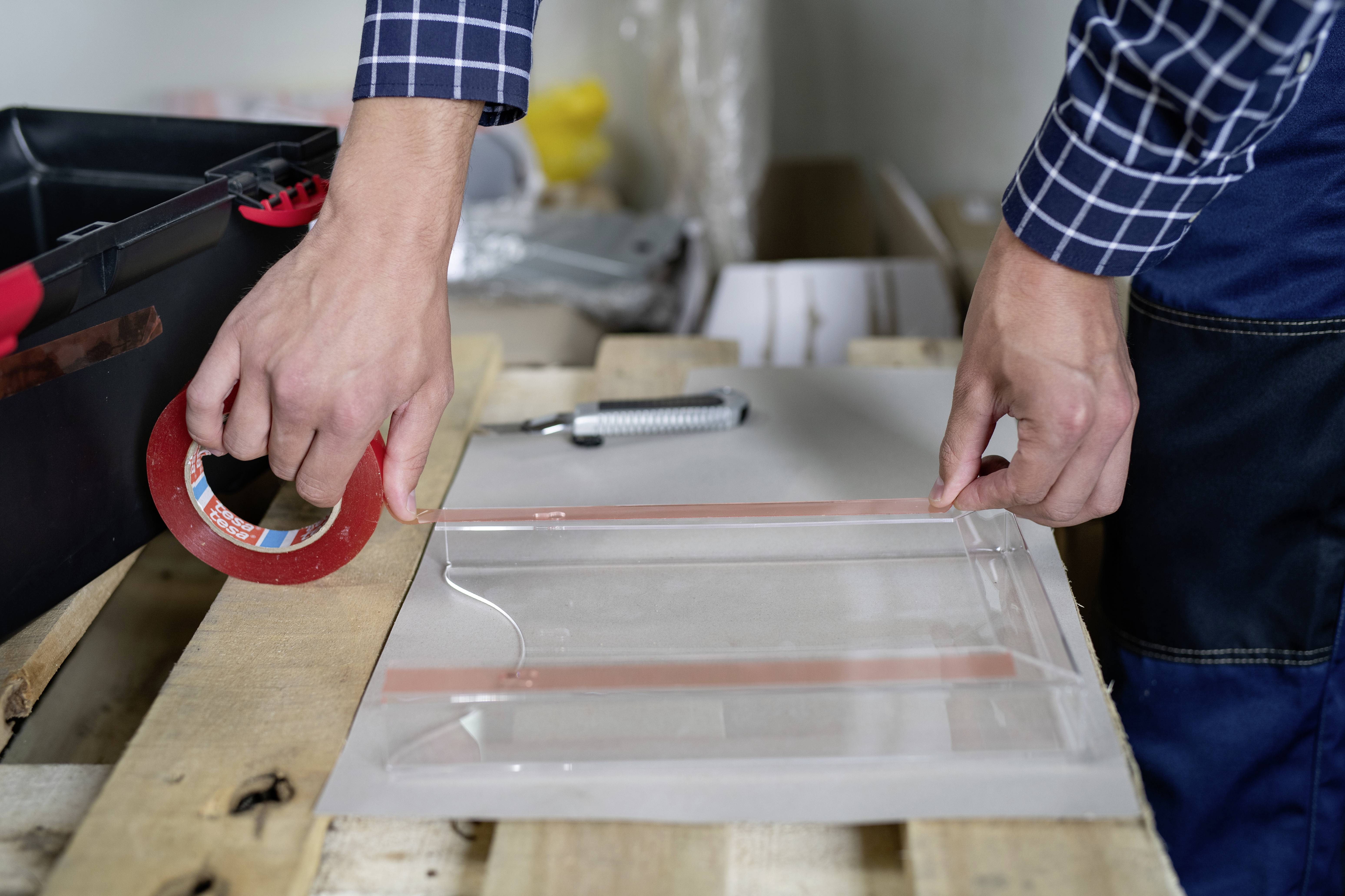 Eine Person verwendet rotes Klebeband, um eine Plastikhülle auf einer Holzpalette zu sichern. Eine Toolbox und Werkzeuge liegen daneben.