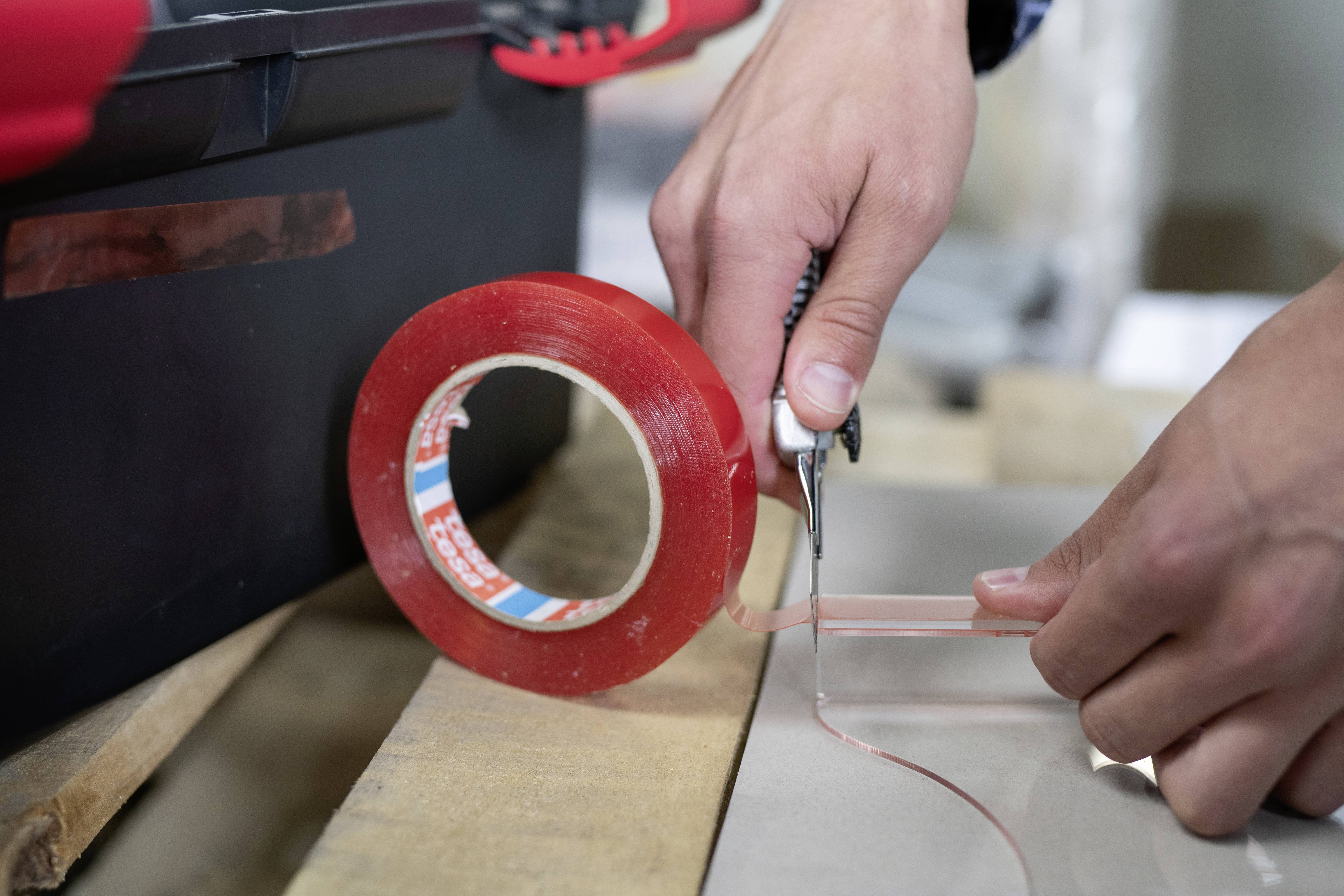 Person schneidet rotes Klebeband mit einem Messer auf einem Holztisch. Rettungsrolle liegt neben einer dunklen Box. Hände sind fokussiert.